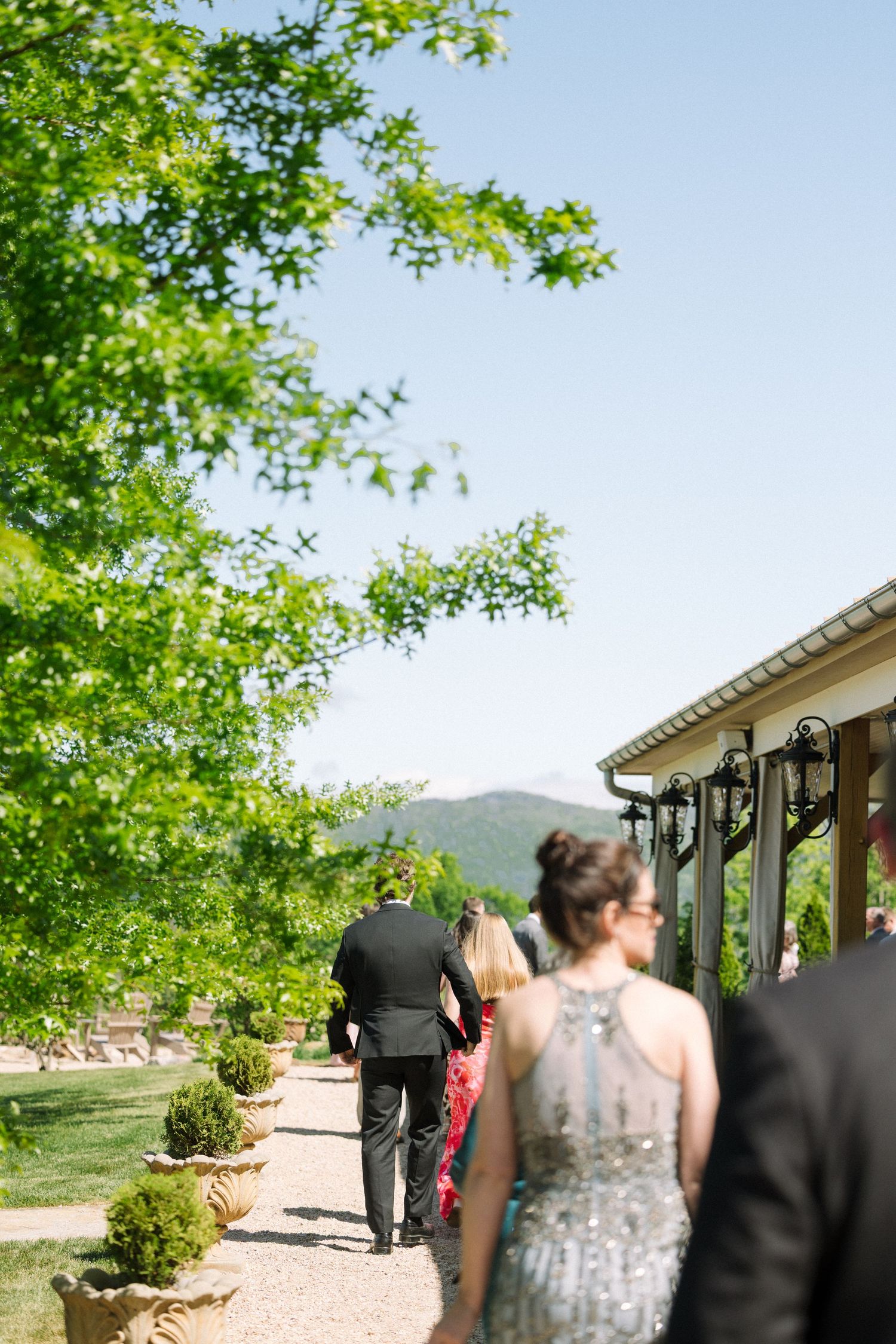 Formally dressed wedding guests walking along a gravel path lined with topiaries beside a rustic mountain venue.