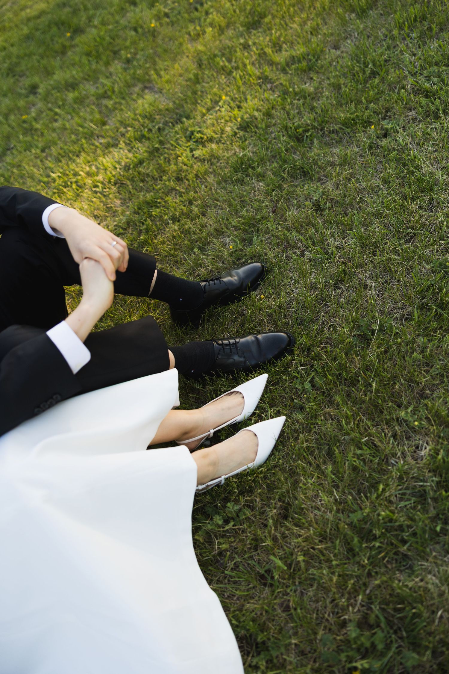 Overhead view of a couple holding hands while sitting on green grass, wearing wedding attire.