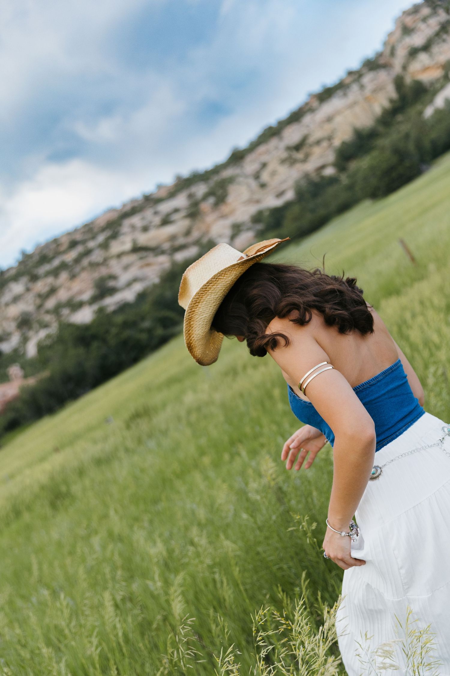 Woman in blue top and cowboy hat bends forward in a lush green mountain meadow.