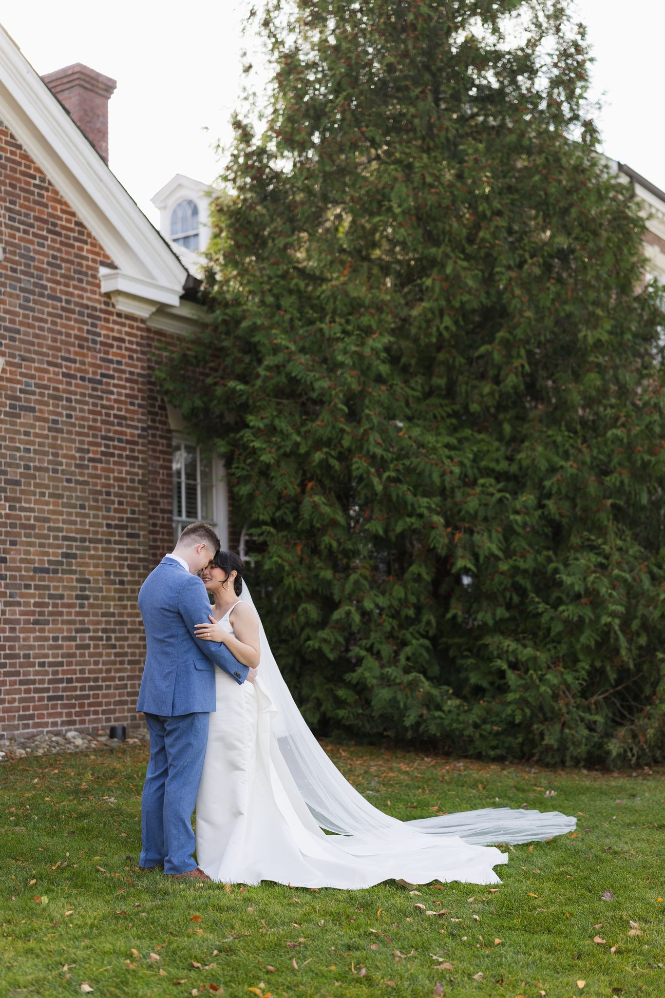 Bride and groom embrace on lawn beside brick building, long wedding veil trailing on grass.