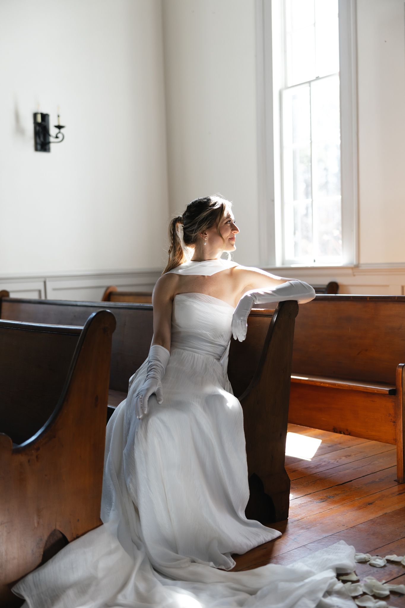 Bride in strapless white gown and gloves sitting on church pew gazing out sunlit window.