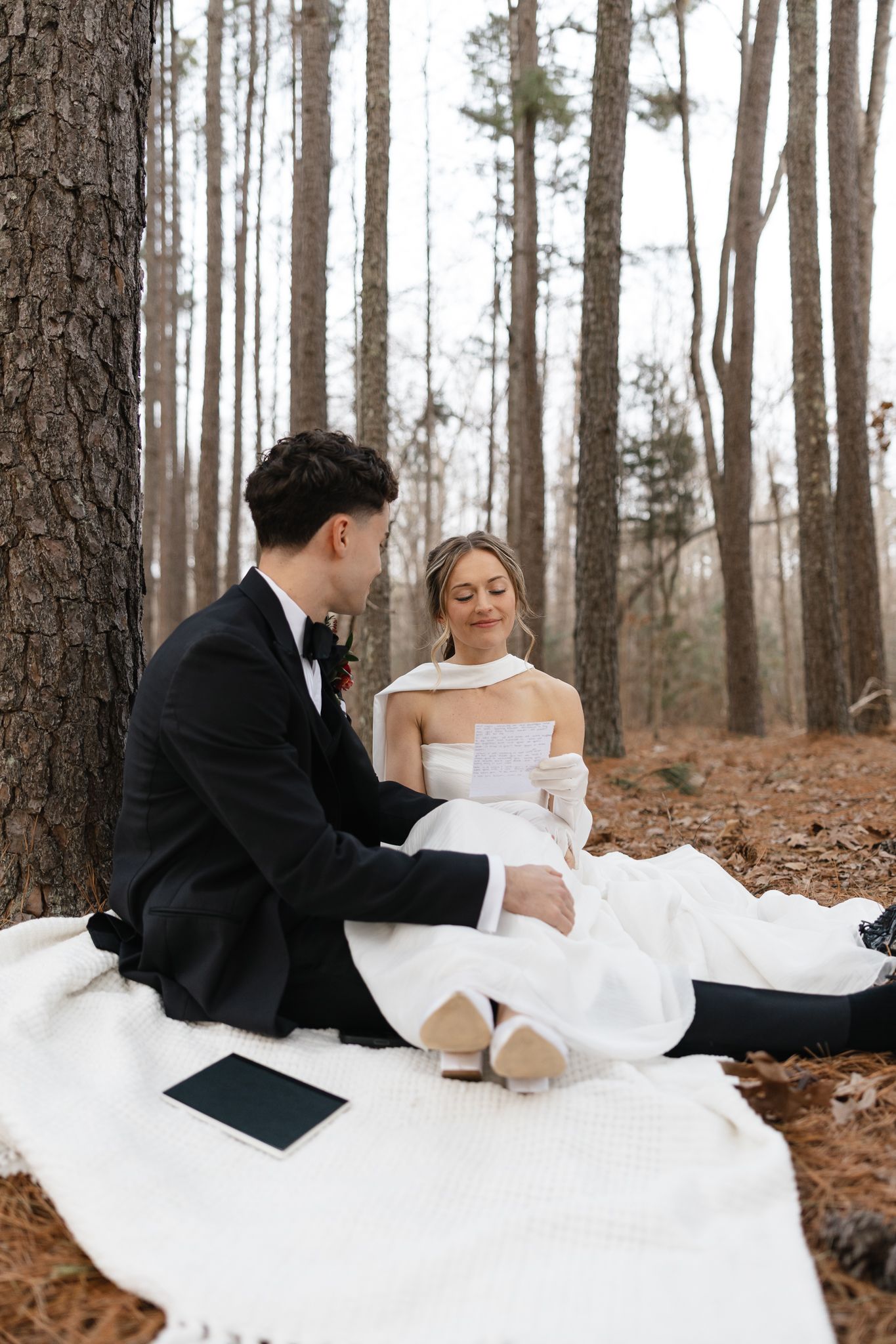 Bride and groom sit on a white blanket in a pine forest, exchanging heartfelt wedding vow letters.