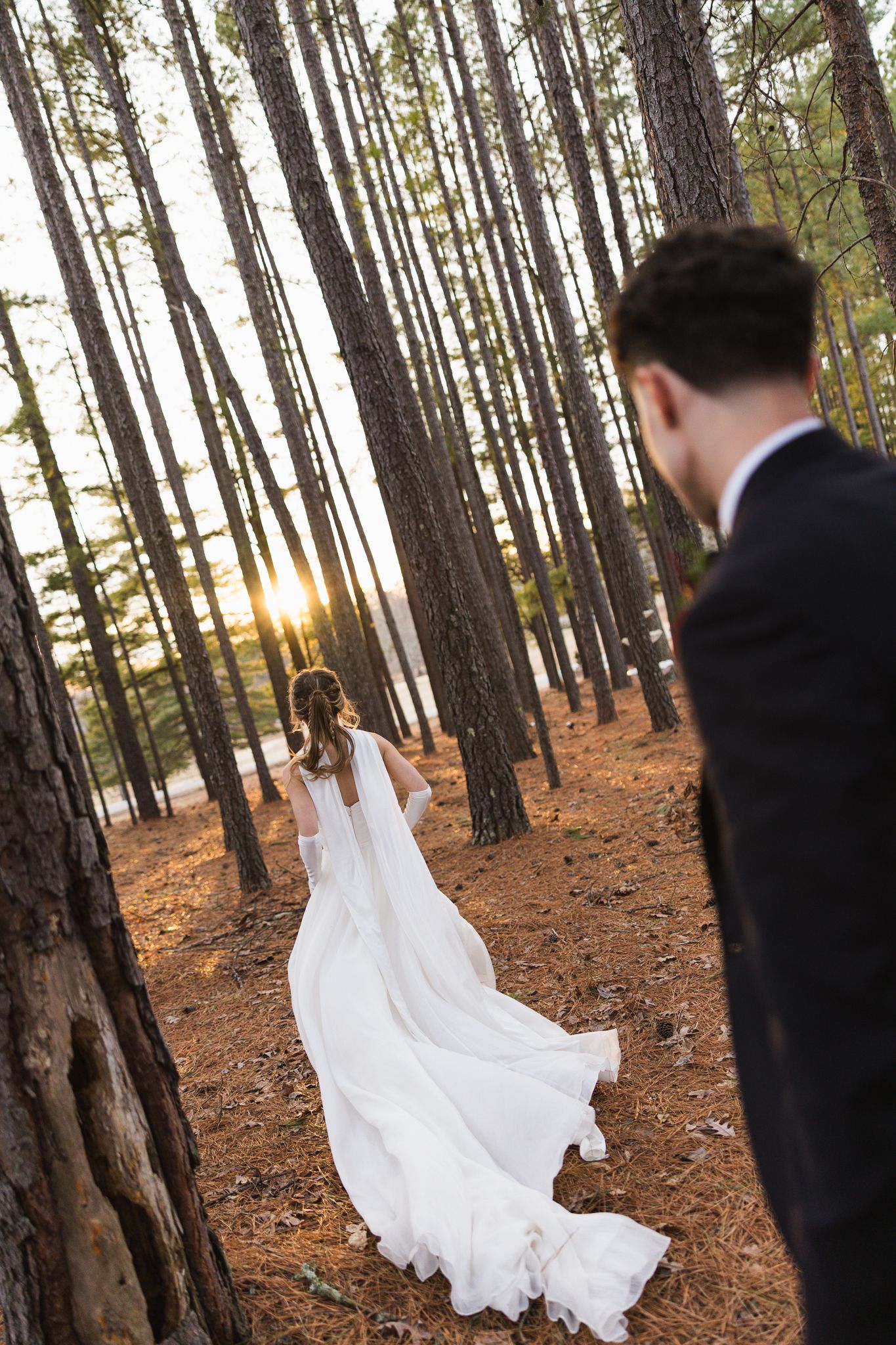 Bride in flowing white gown walks through pine forest at golden sunset during wedding photo session.