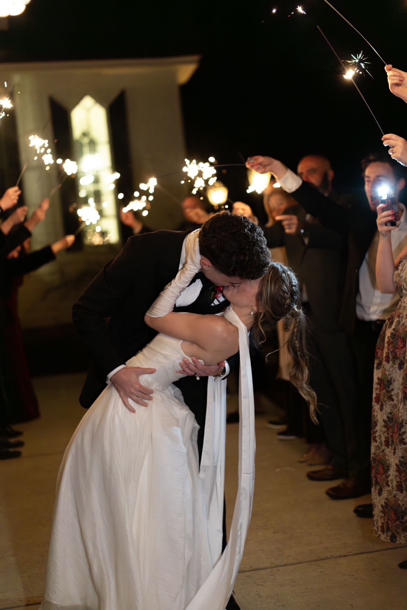 Bride and groom share a dramatic dip kiss during sparkler send-off surrounded by cheering wedding guests at night.