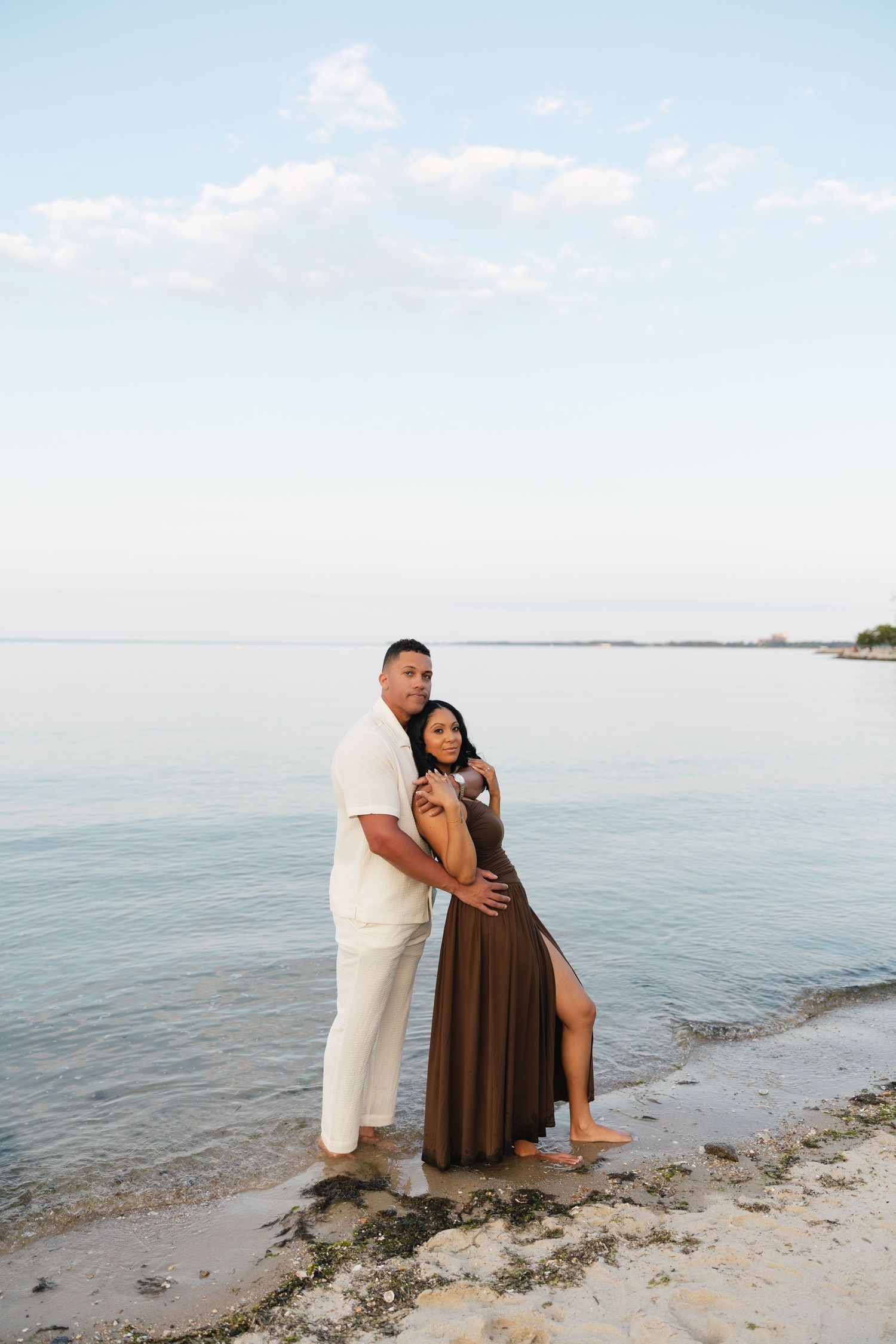 Couple embracing barefoot on sandy beach shore, woman in brown dress and man in white outfit by calm ocean.
