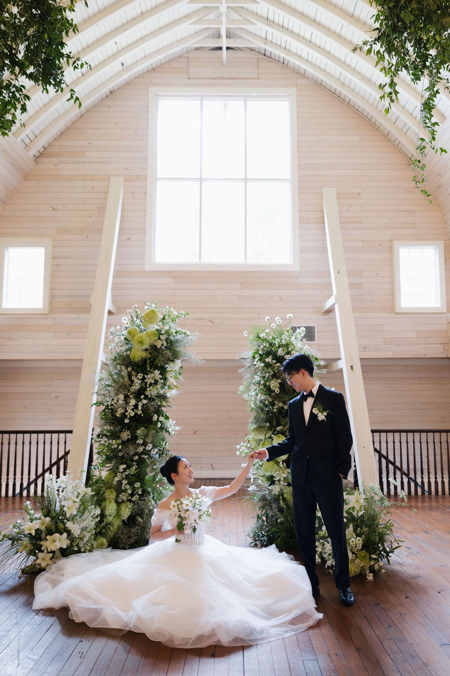 Bride in white gown sits on wooden floor as groom holds her hand beside lush floral arch in elegant barn venue.