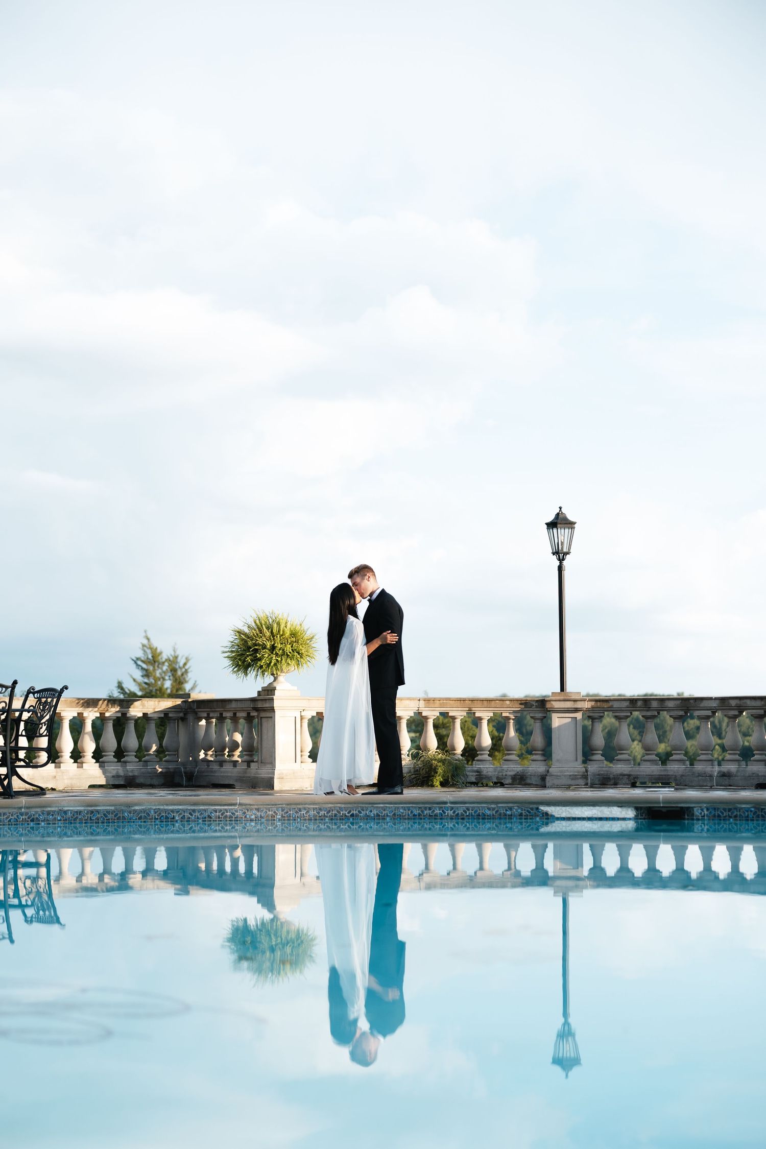 Bride and groom kissing by elegant poolside balustrade, their reflection mirrored in calm blue water at a luxury venue.