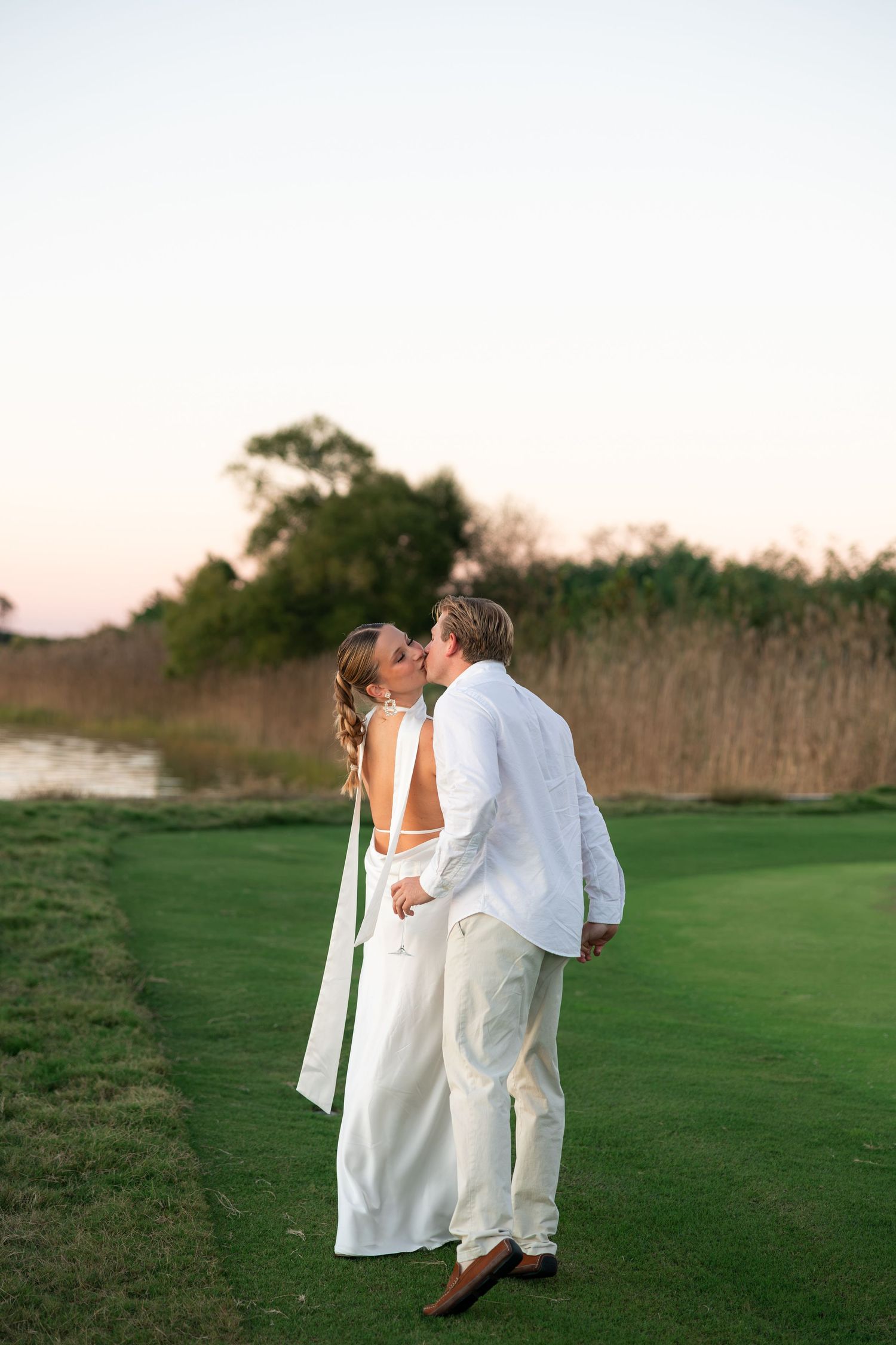 Bride and groom sharing a kiss on a golf course at sunset, both dressed in elegant white wedding attire.