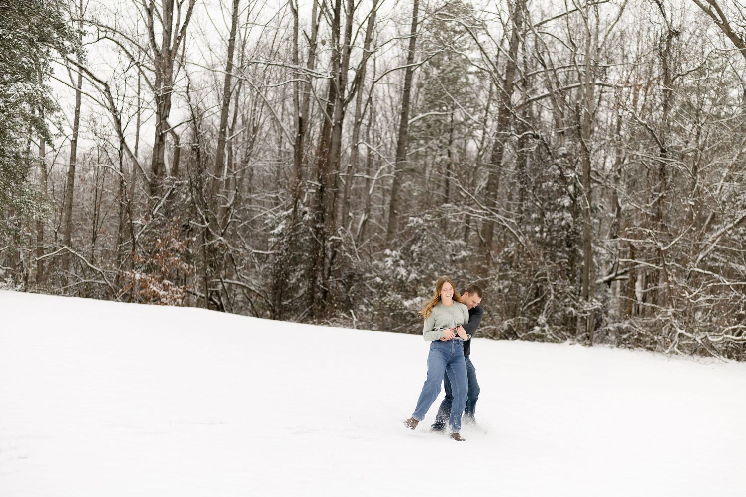 Happy couple playing and embracing on a snowy hill surrounded by snow-covered winter trees.