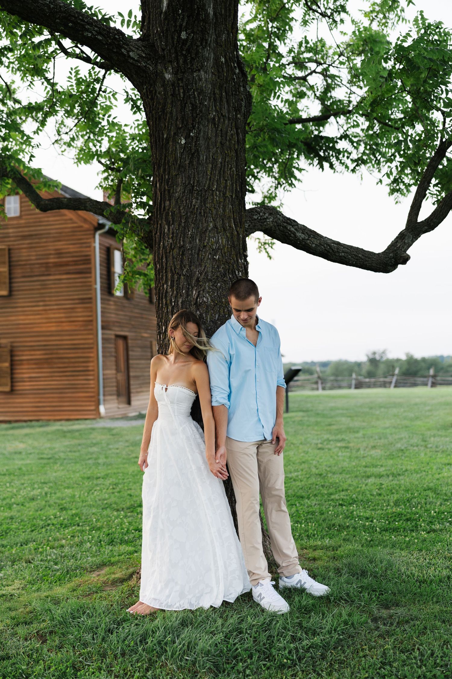 A couple in wedding attire holds hands under a large tree on a green lawn near a wooden building.