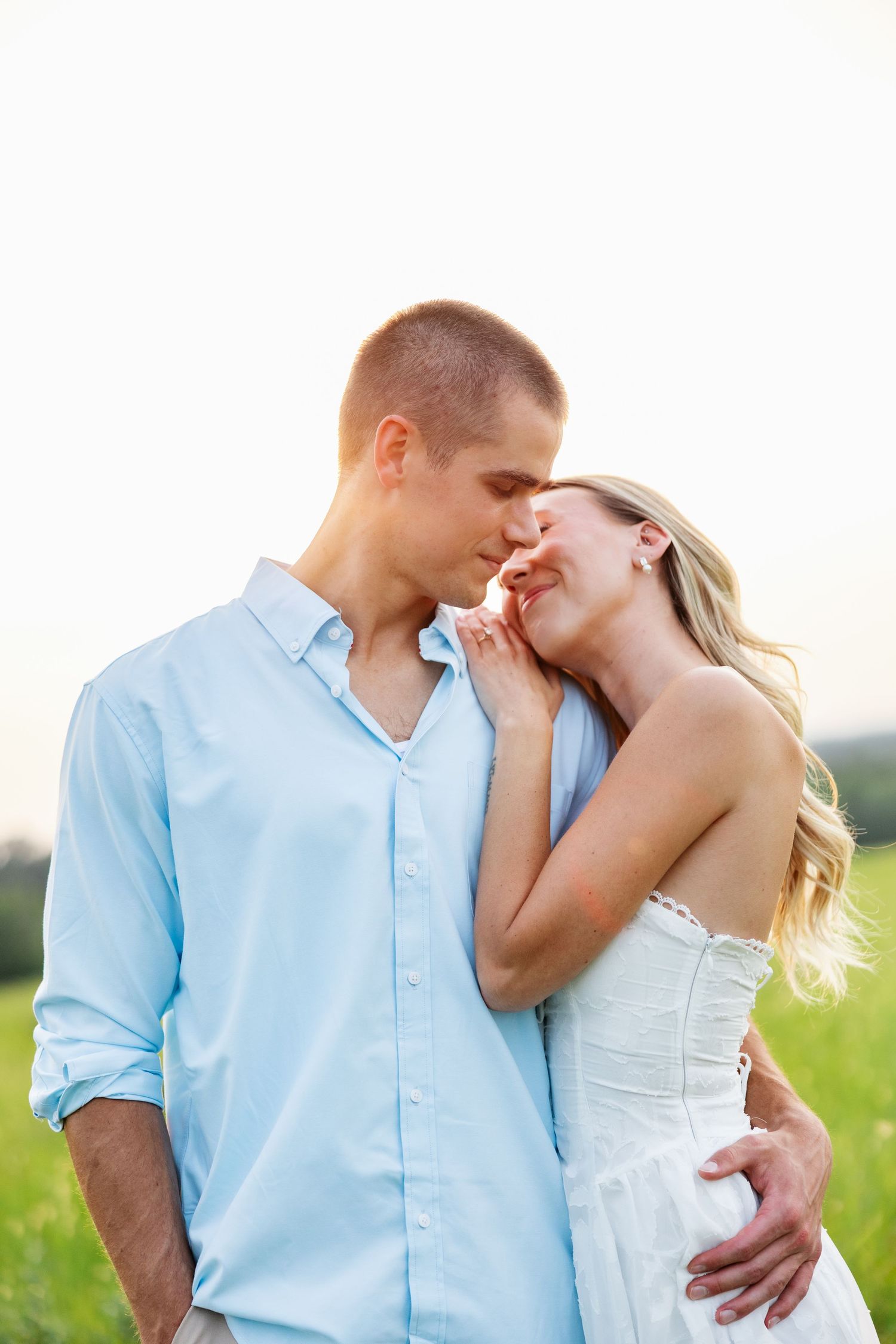 Romantic couple sharing a tender kiss in a sunny green field, man in blue shirt embracing woman in white dress.