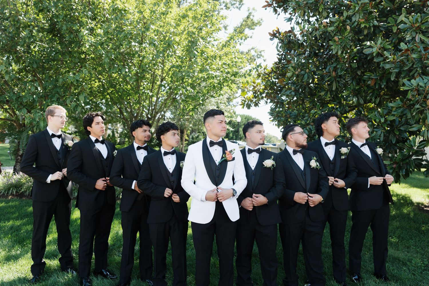 Groom in white tuxedo jacket stands with groomsmen in black tuxedos and bow ties outdoors on a sunny day.