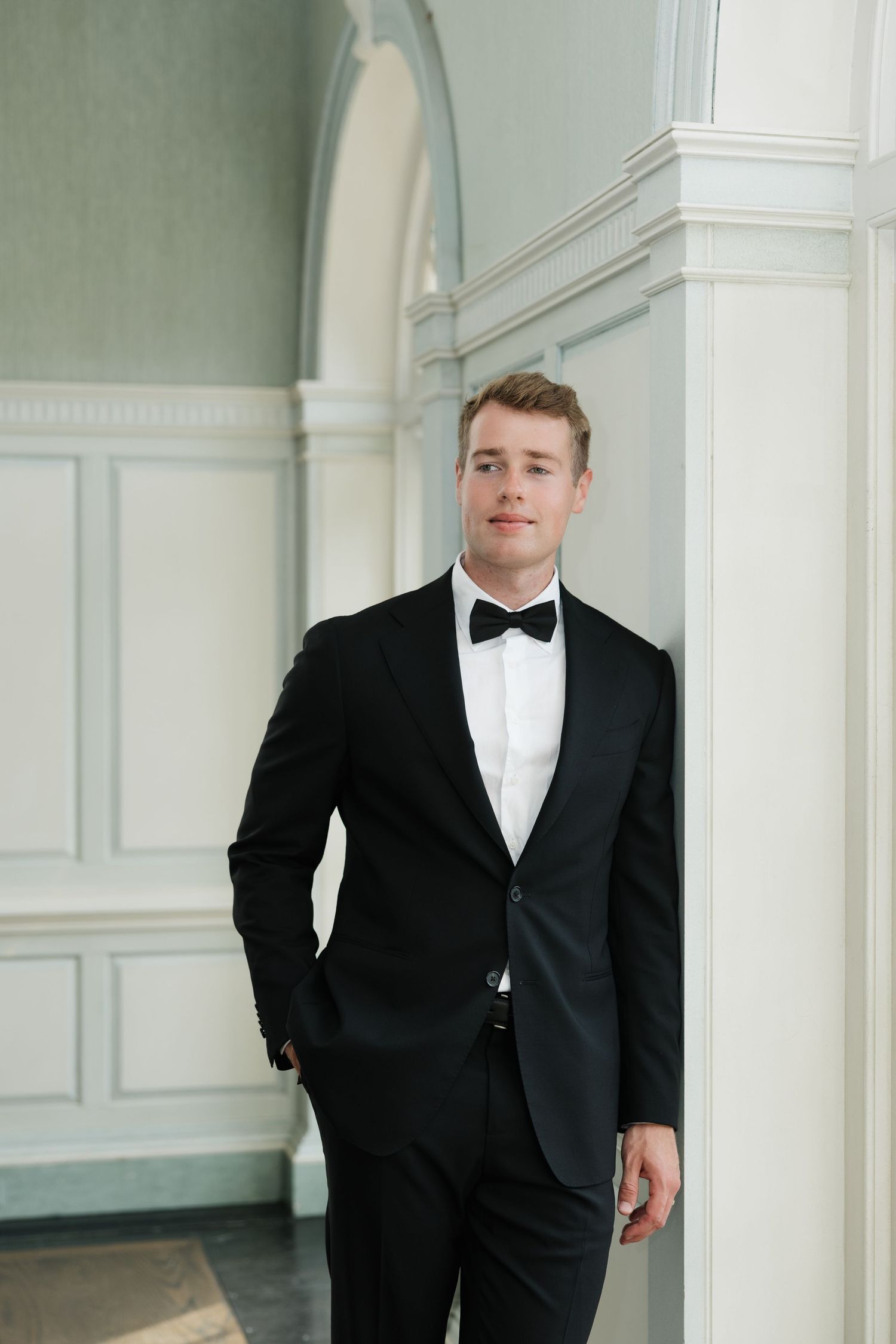 Young man in black tuxedo and bow tie leaning against white column in elegant interior setting.