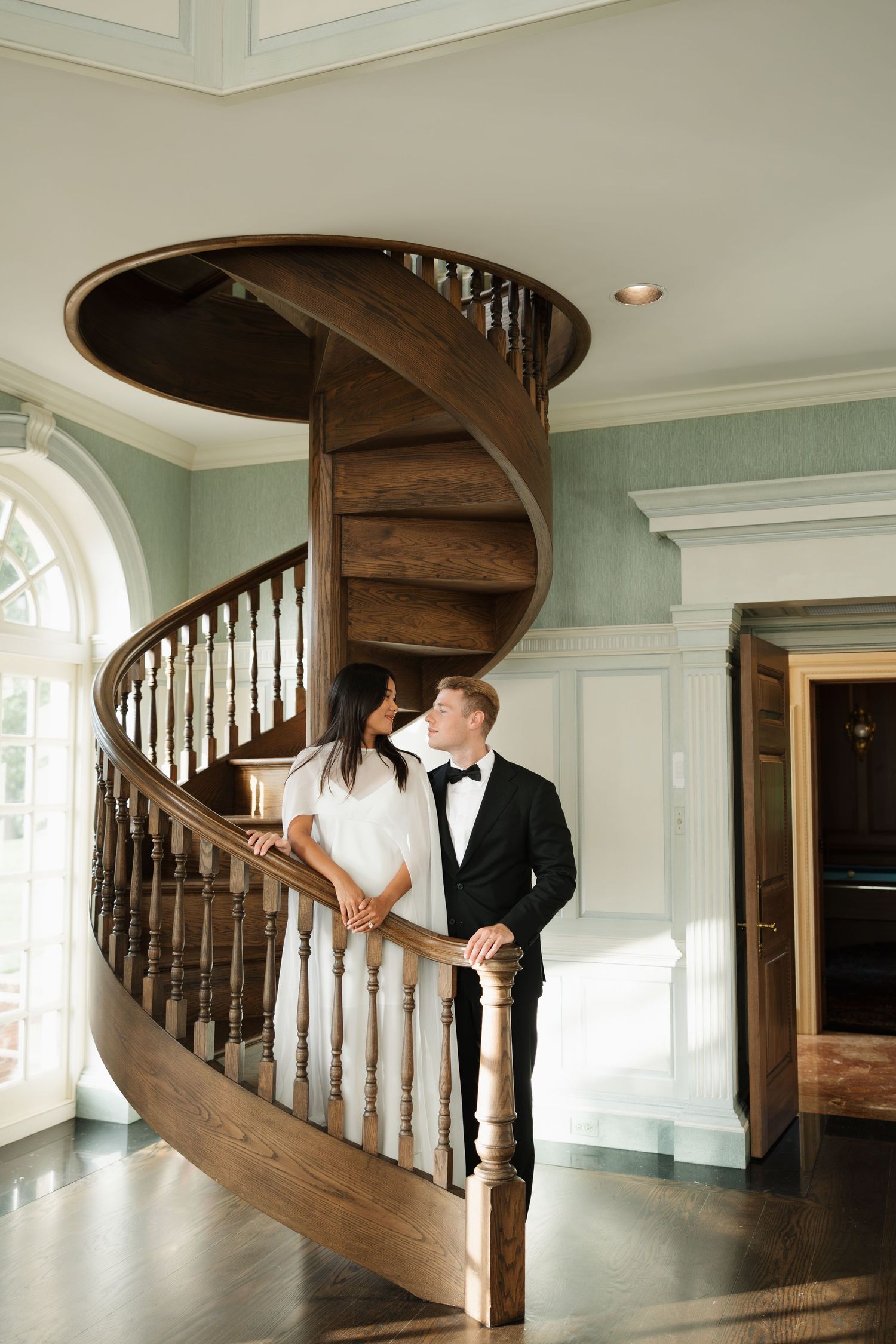 Couple in wedding attire sharing a romantic moment on an elegant spiral staircase in a grand estate.