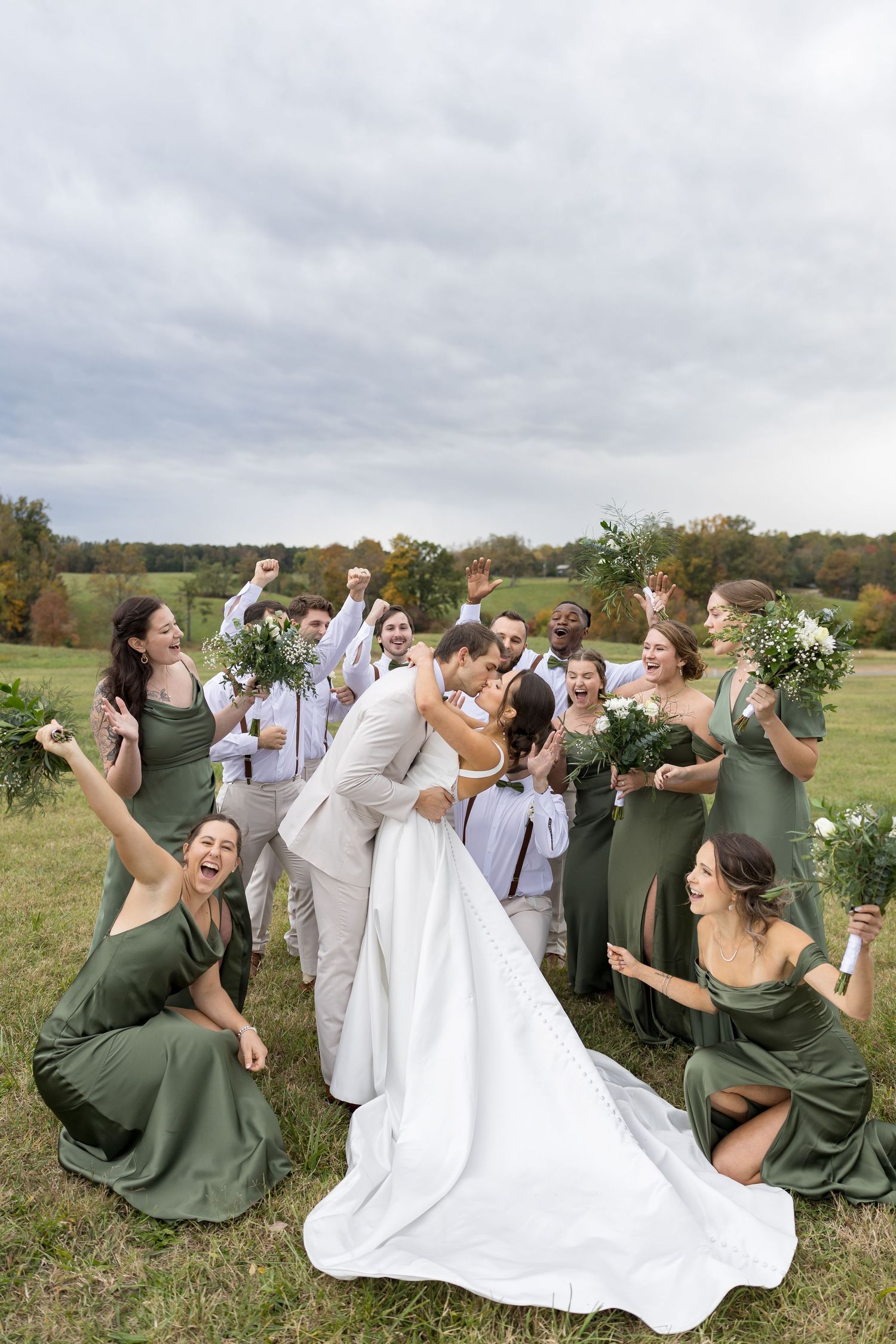 Wedding party celebrates as bride and groom kiss in green field, bridesmaids in sage dresses cheering.