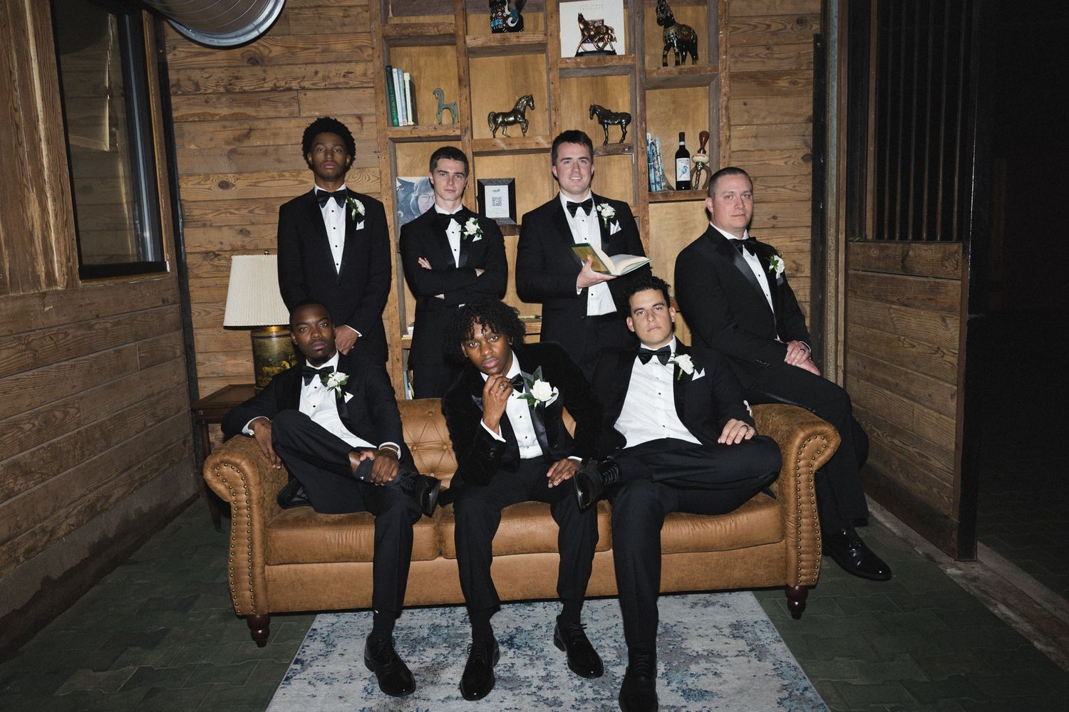 Groomsmen in black tuxedos posing casually on leather sofa in rustic wood-paneled room.