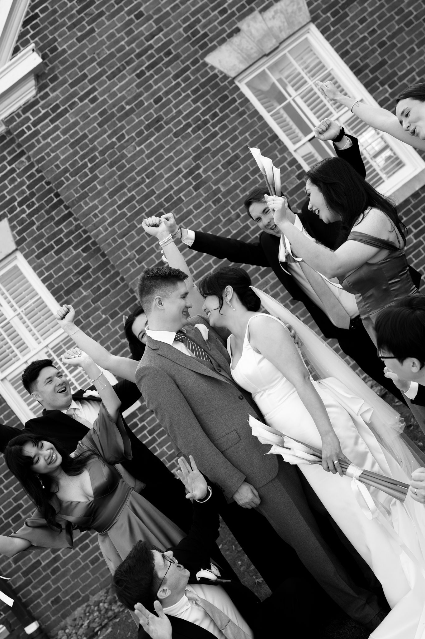 Bride and groom share kiss surrounded by cheering wedding guests outside brick building, black and white.