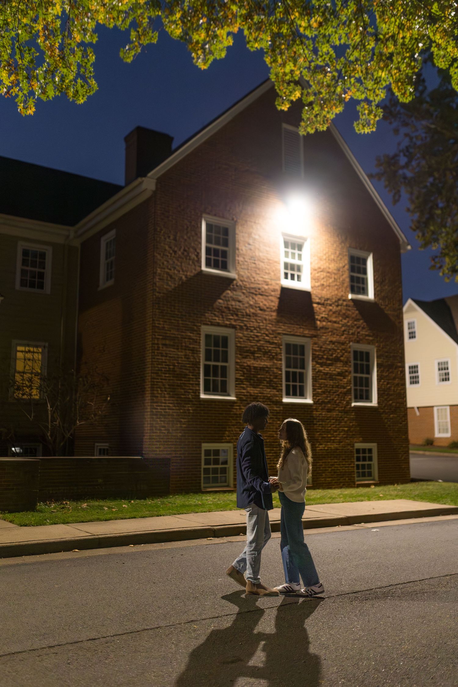 A couple holds hands walking along a street at night, illuminated by a bright light on a brick building.