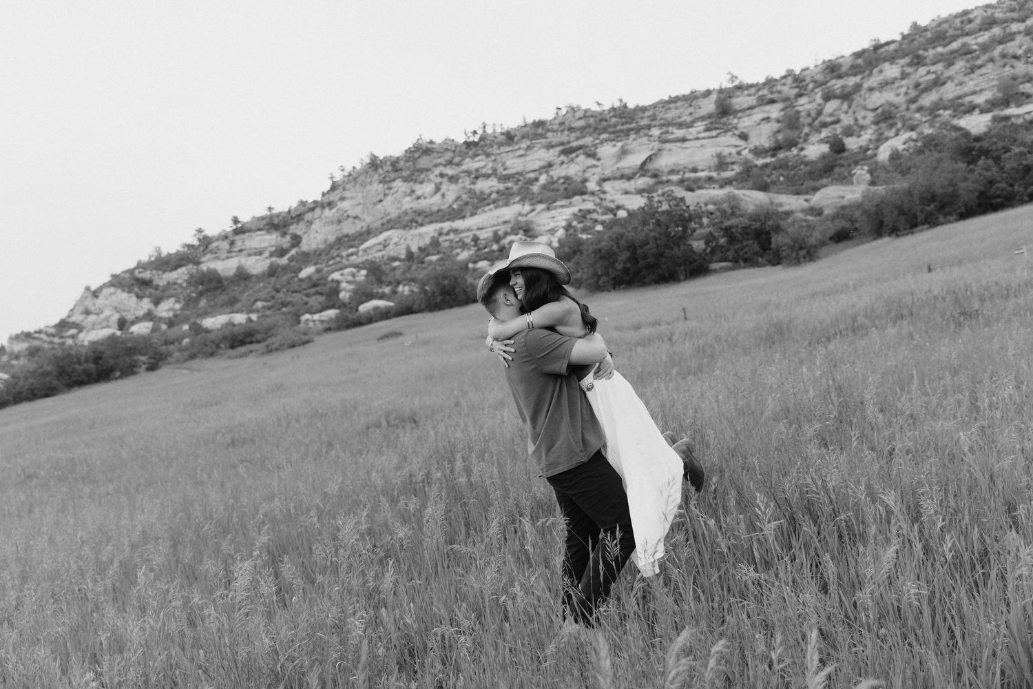A couple shares a romantic embrace in a vast green field with a rocky hillside in black and white.