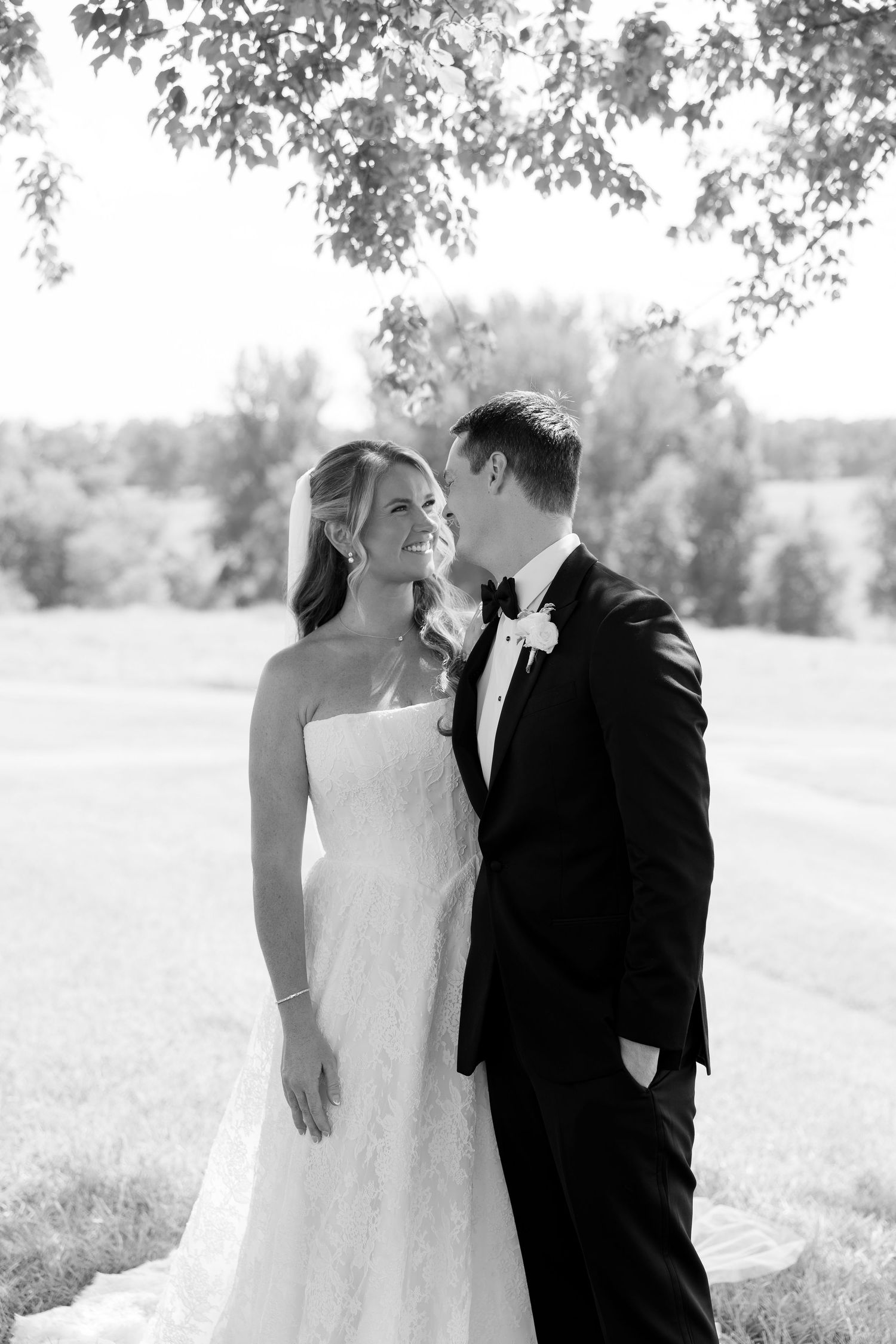 Black and white wedding photo of smiling bride in strapless lace gown with groom in tuxedo outdoors.
