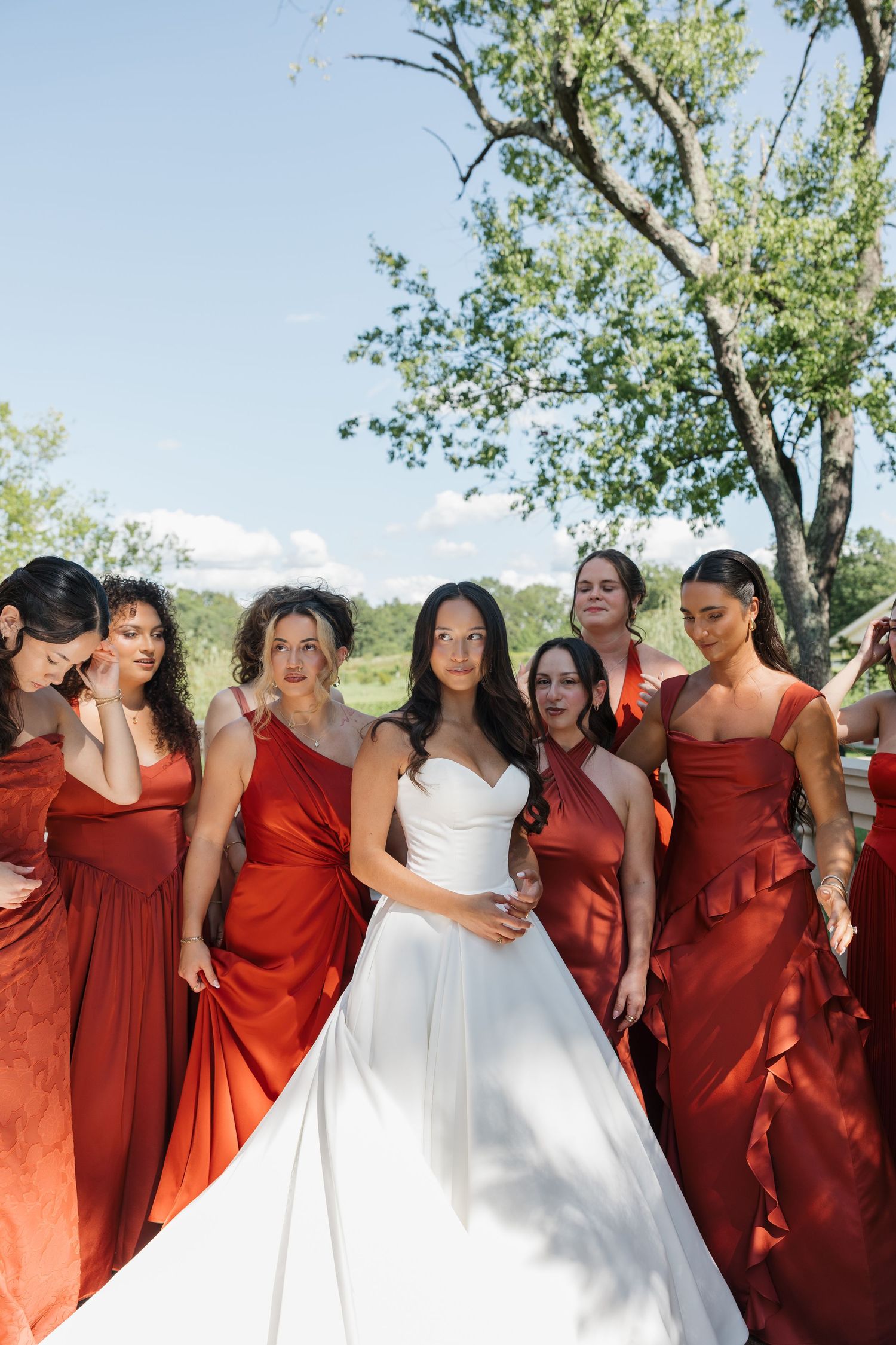 Bride in white gown surrounded by bridesmaids in red satin dresses outdoors on a sunny day.