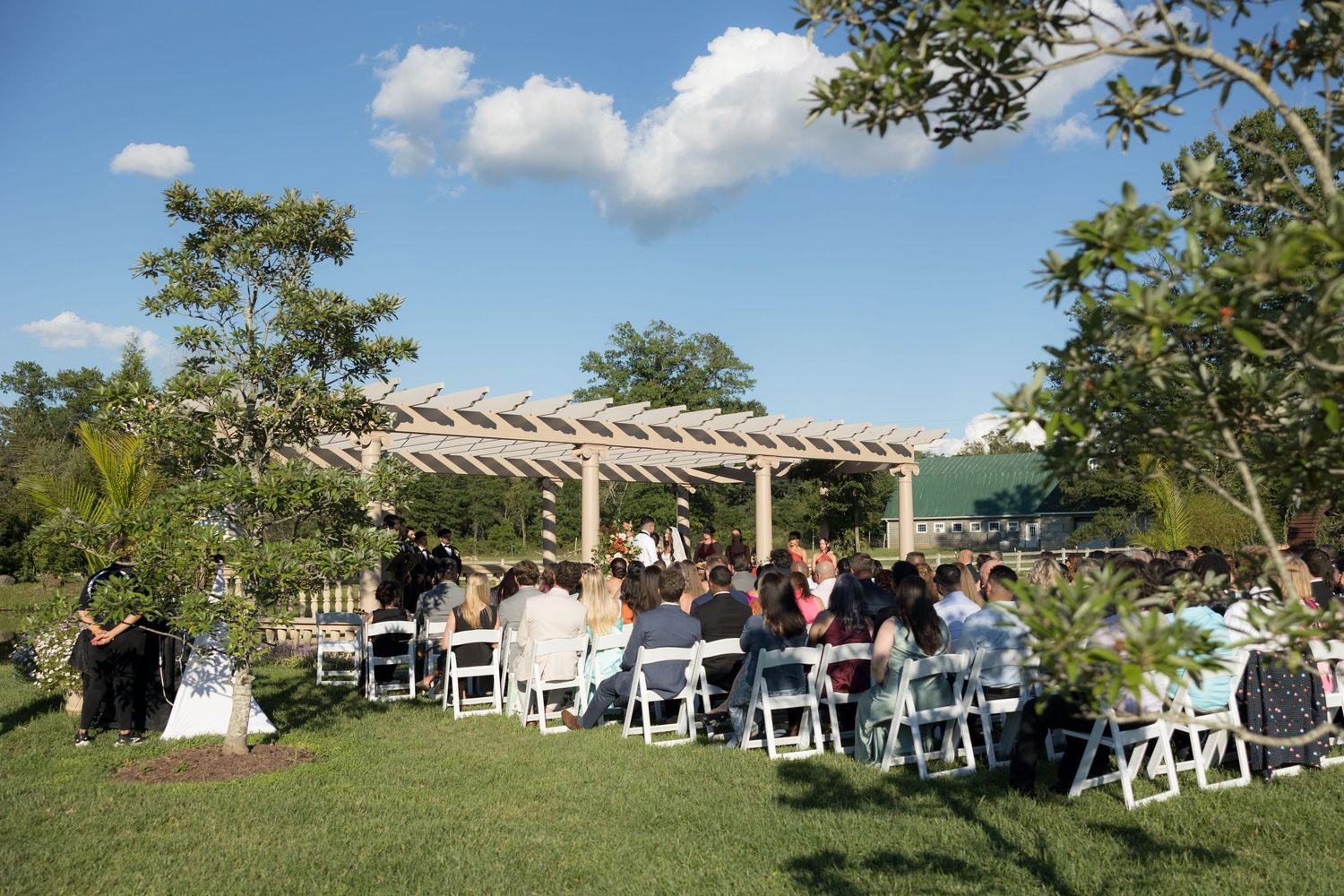 Outdoor wedding ceremony on green lawn with white chairs and wooden pergola under blue sky.