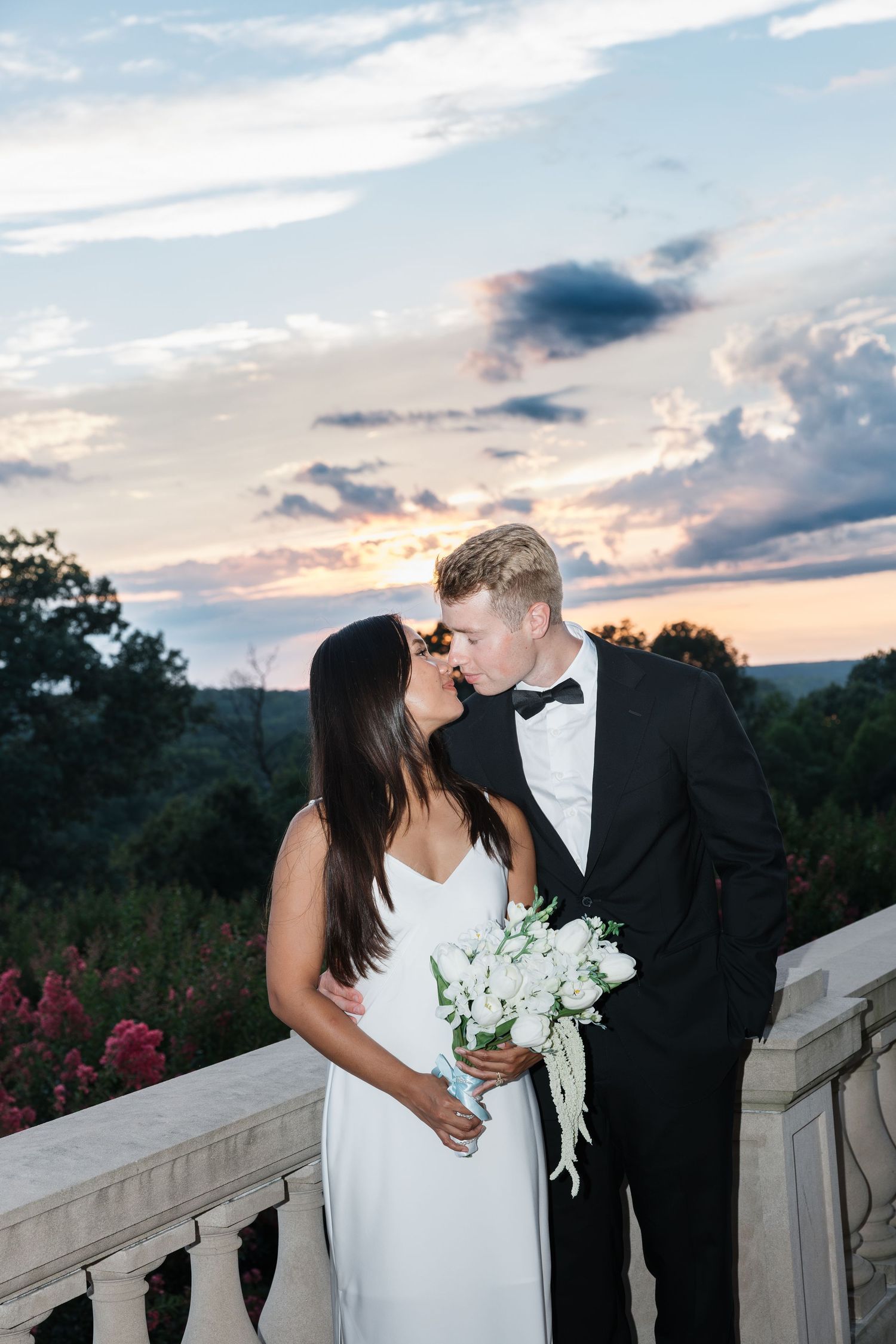 Newlyweds sharing a kiss at sunset on a balcony, bride holding white bouquet, groom in black tuxedo.