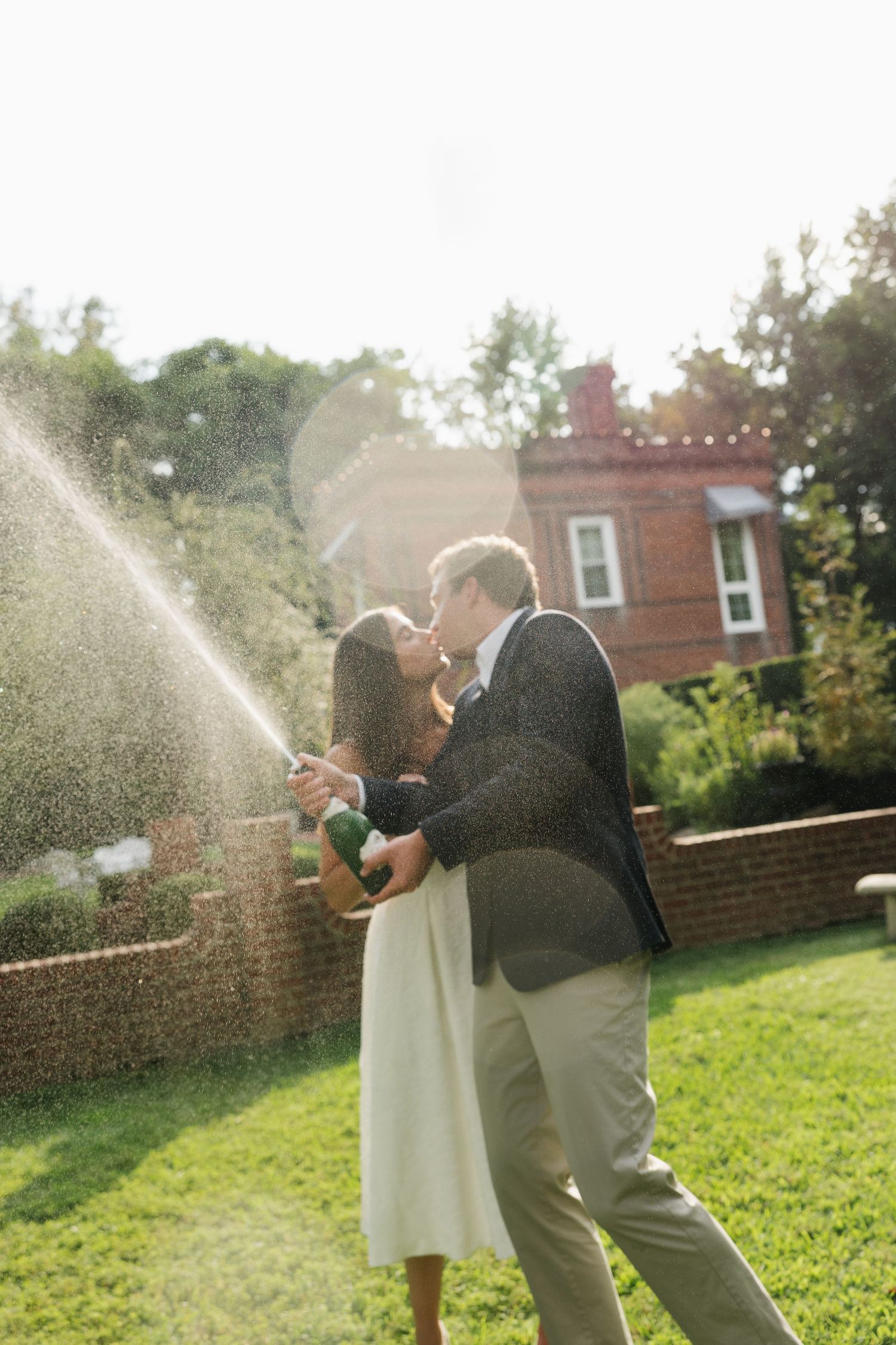 Couple kissing while popping champagne bottle outdoors on green lawn with brick building in background.