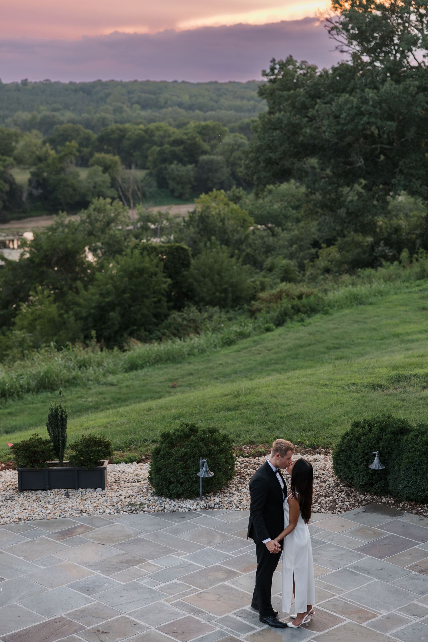 Couple sharing a romantic moment on a stone patio at sunset with lush green hills and dramatic sky backdrop.