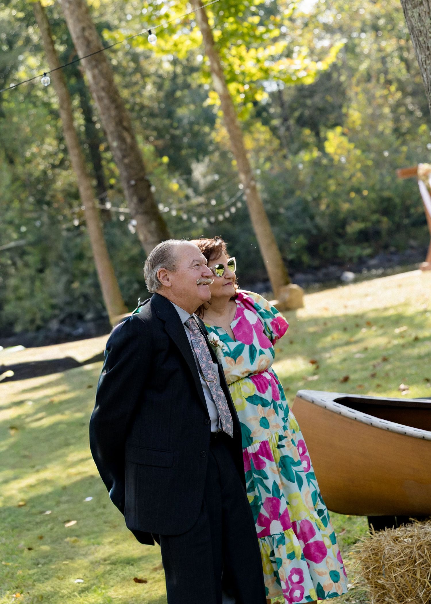 Elderly couple in formal attire standing near hay bales and wooden canoe at outdoor garden wedding celebration.