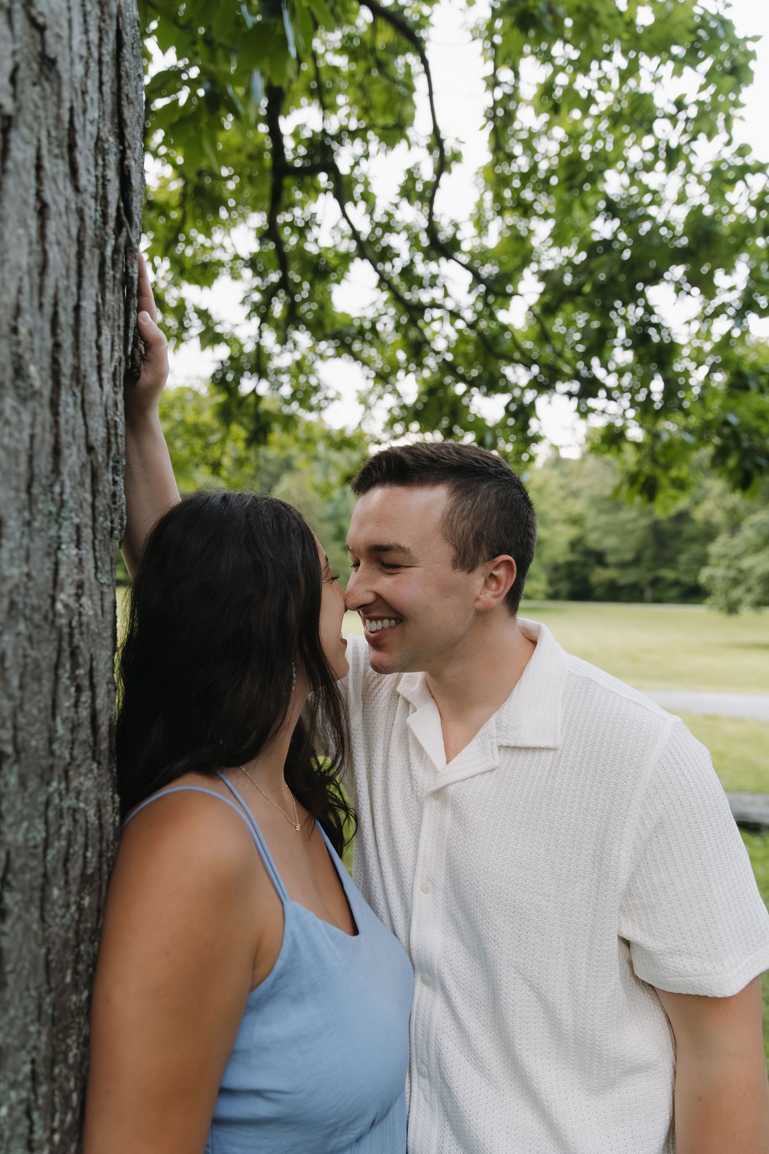 Laughing couple leaning against tree trunk in green park setting.