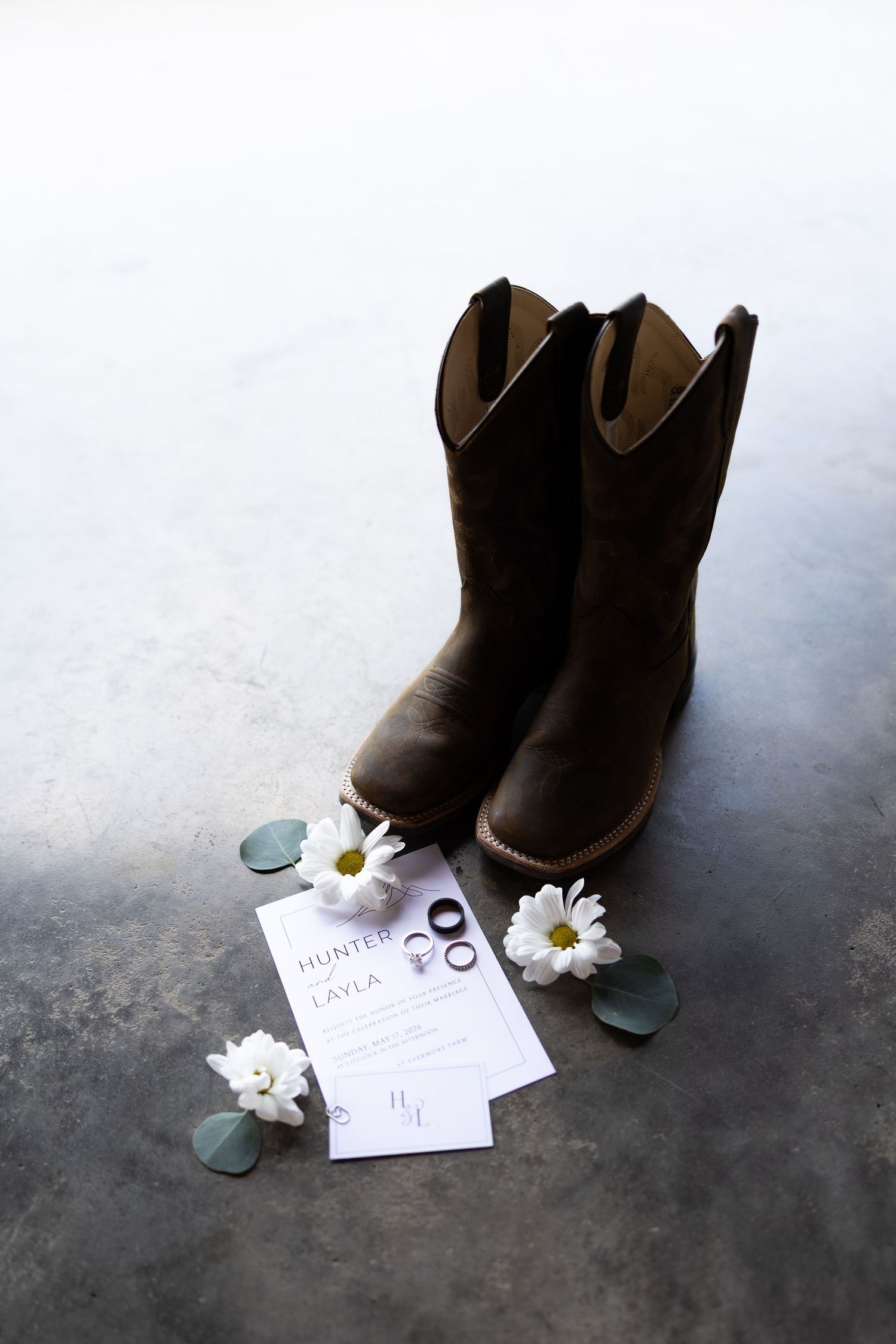 Rustic wedding flat lay with brown cowboy boots, daisy flowers, wedding rings, and invitation cards on gray surface.