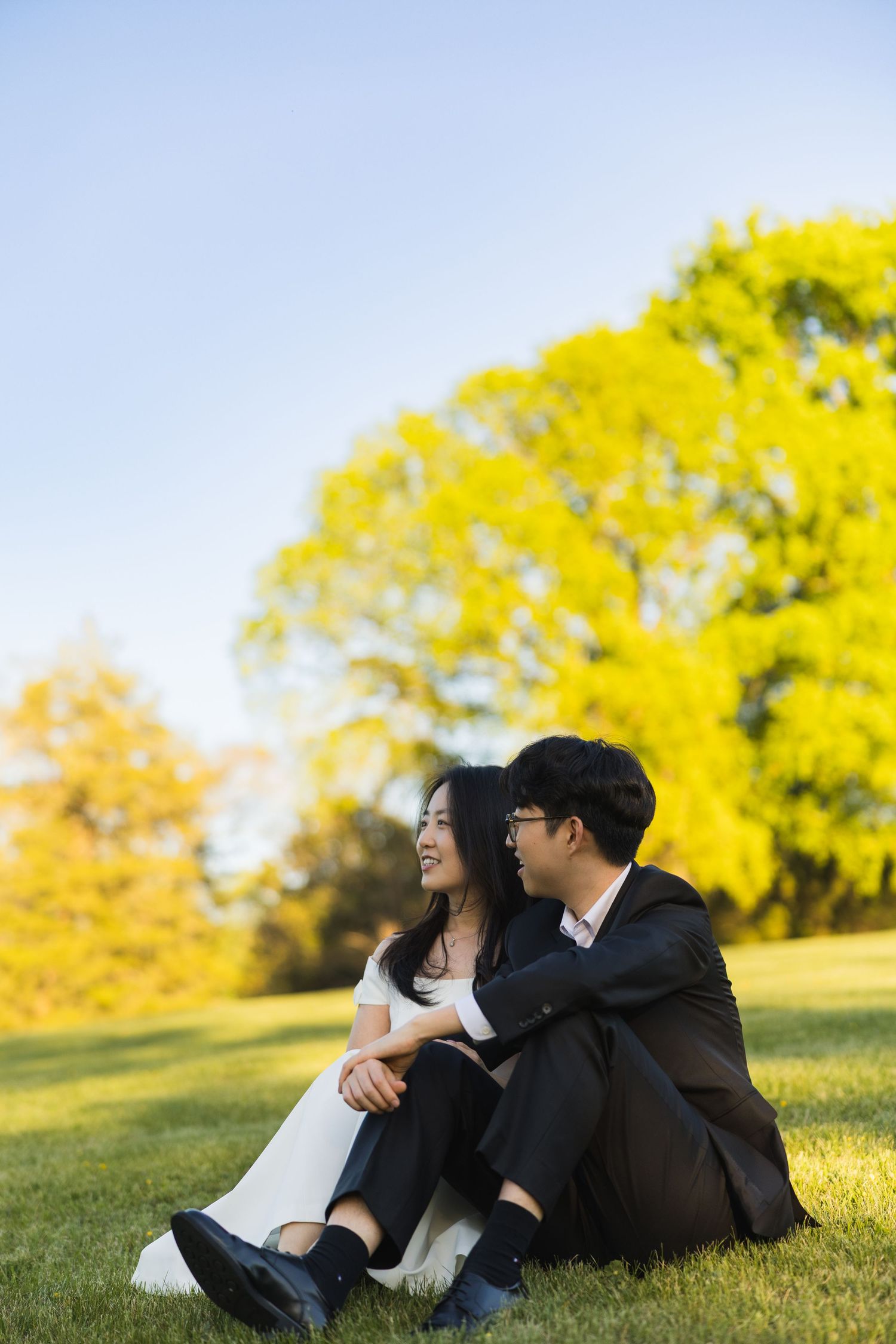 Couple sitting on grass in wedding attire, smiling at each other with vibrant yellow trees behind.