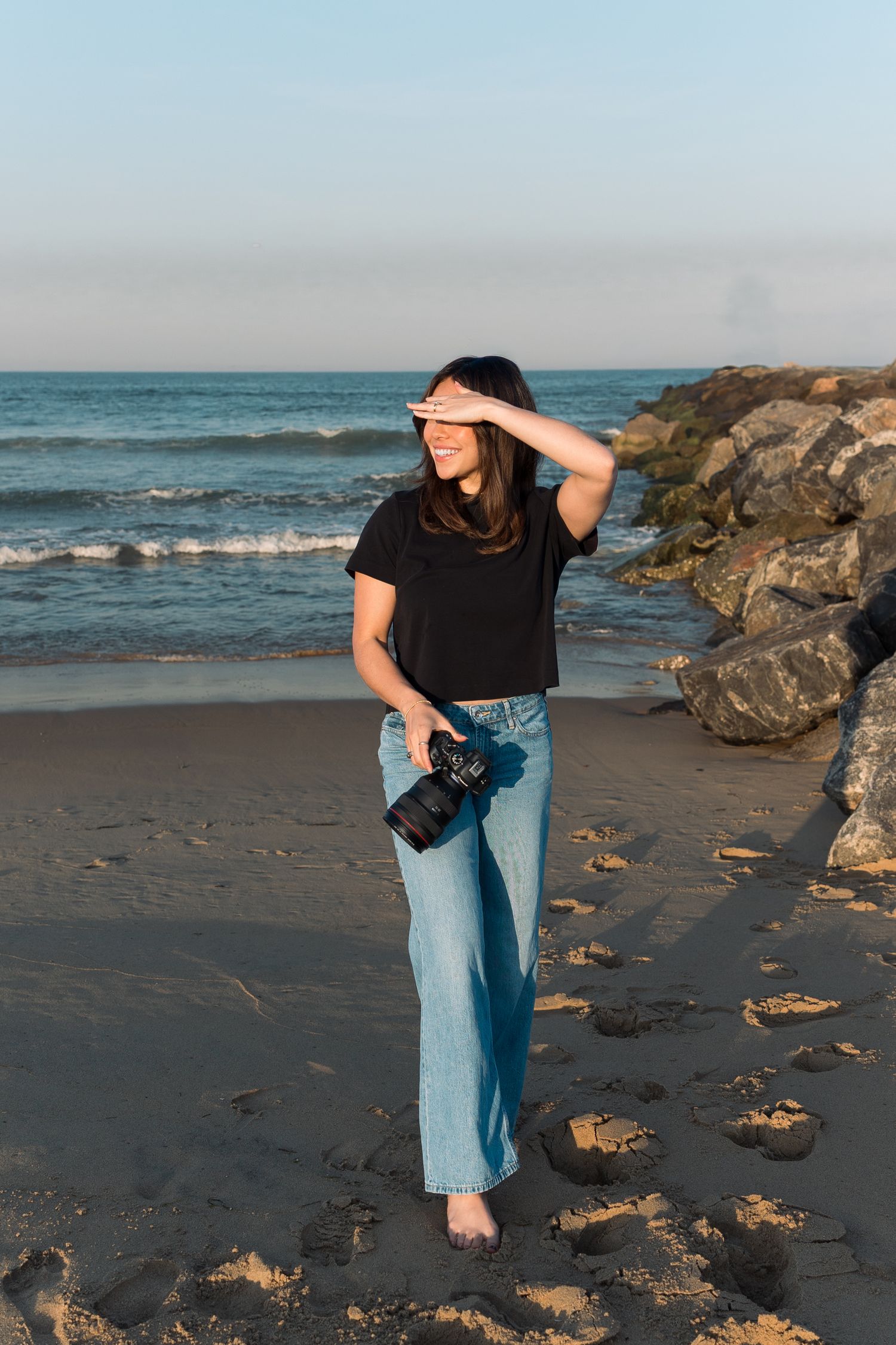 Smiling woman in black crop top and jeans holds camera while shielding eyes from sun on rocky beach at sunset.
