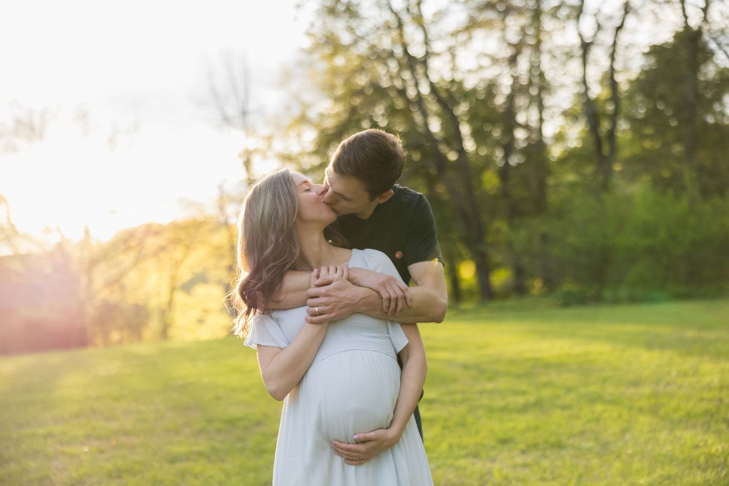 Couple embracing and kissing in sunny green park, woman in white dress with visible baby bump.
