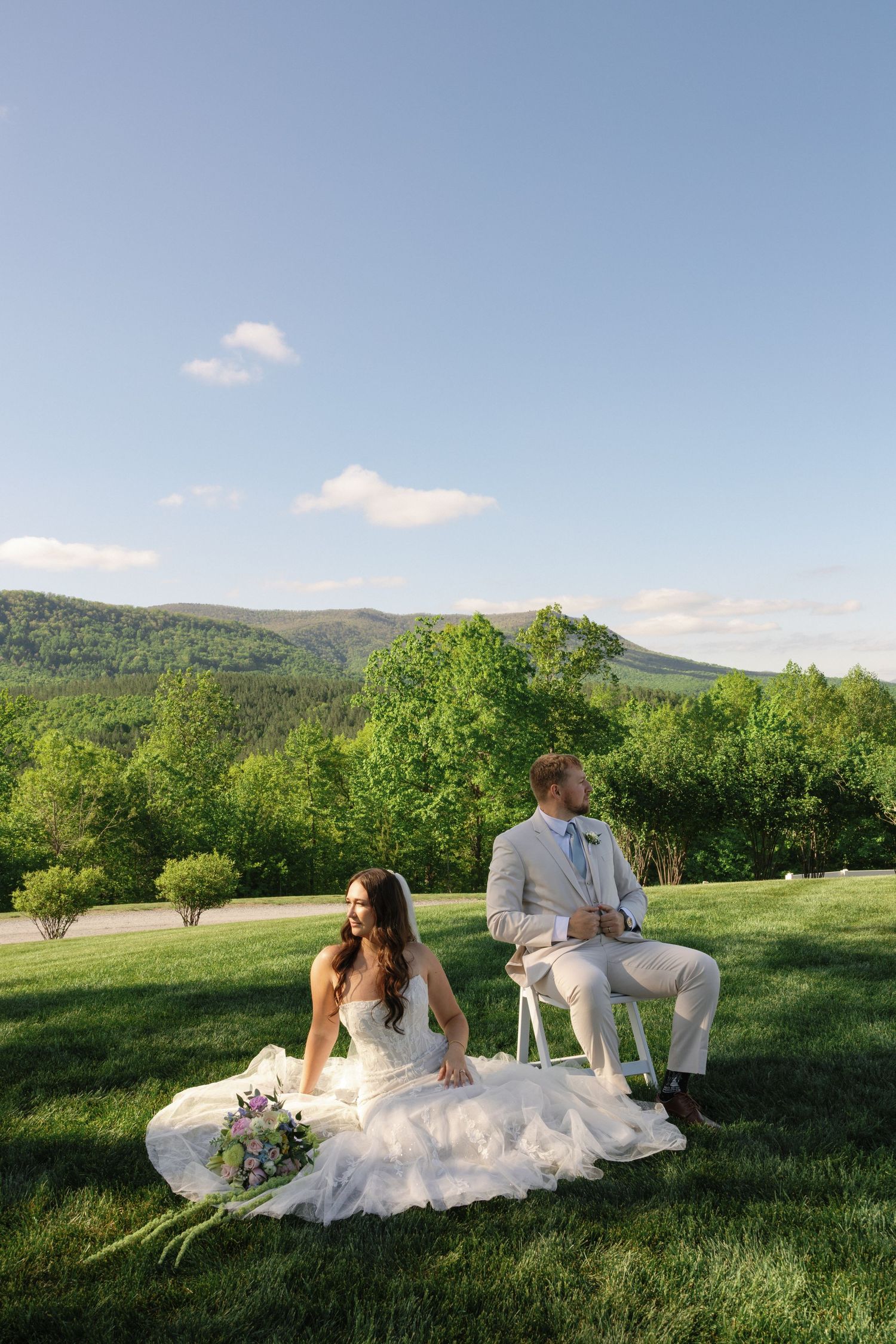 Bride and groom sitting on a lush green lawn with rolling mountain views under a clear blue sky.