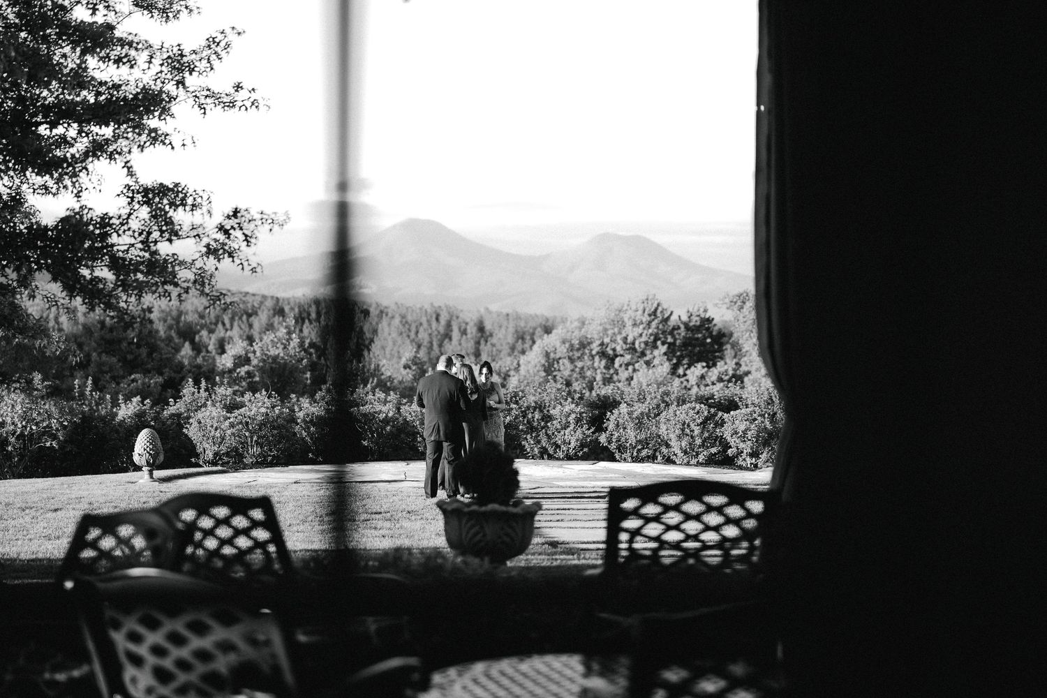 Black and white view through window of couple embracing on porch overlooking scenic mountain landscape.