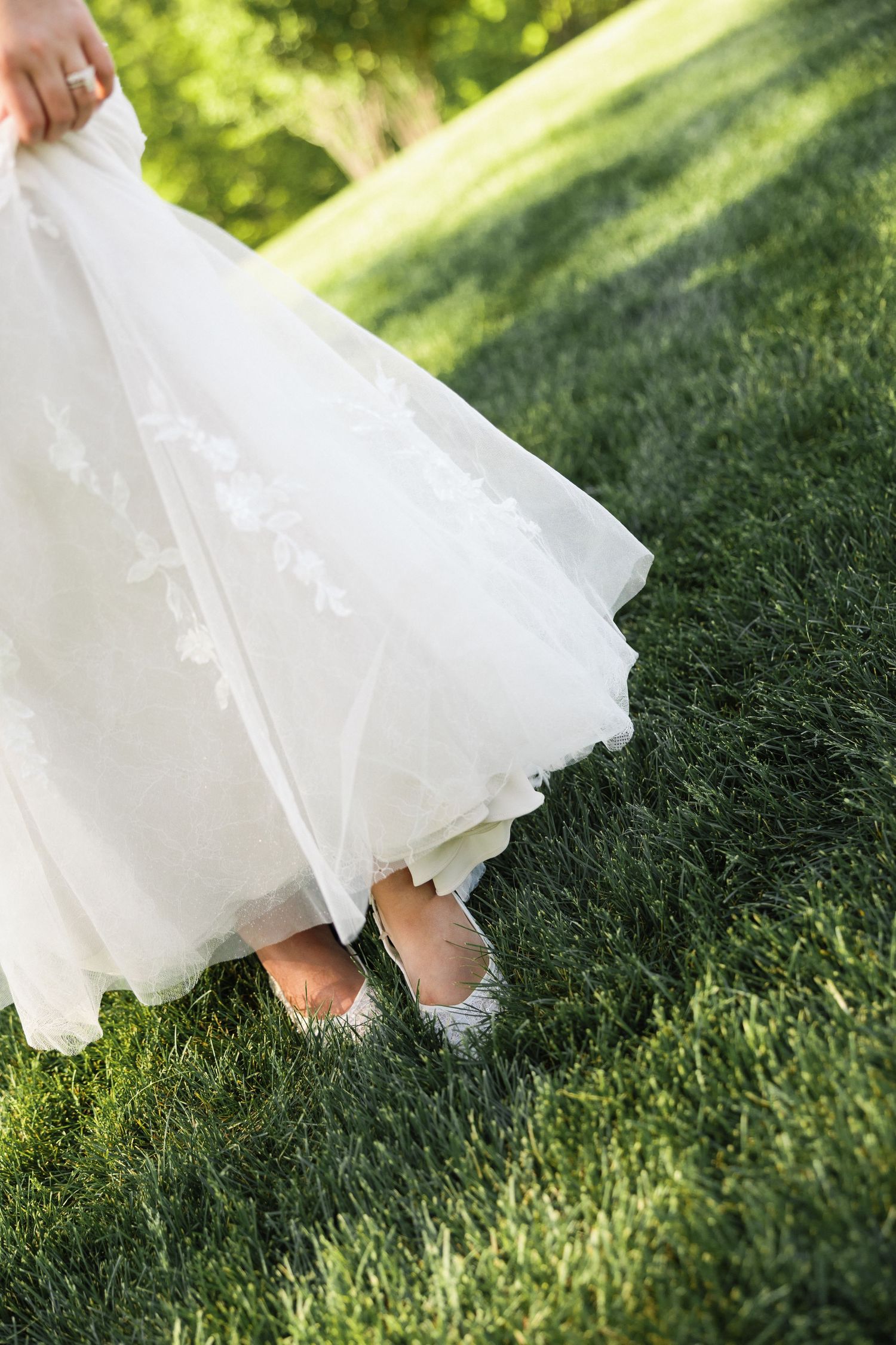 Close-up of bride lifting white lace wedding gown hem revealing silver glitter flat shoes on green grass.