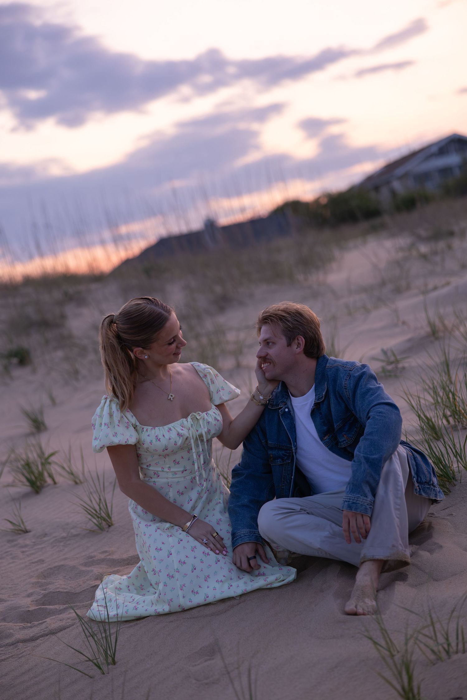 Couple sits on sandy dunes at sunset, woman gently touching mans face romantically.