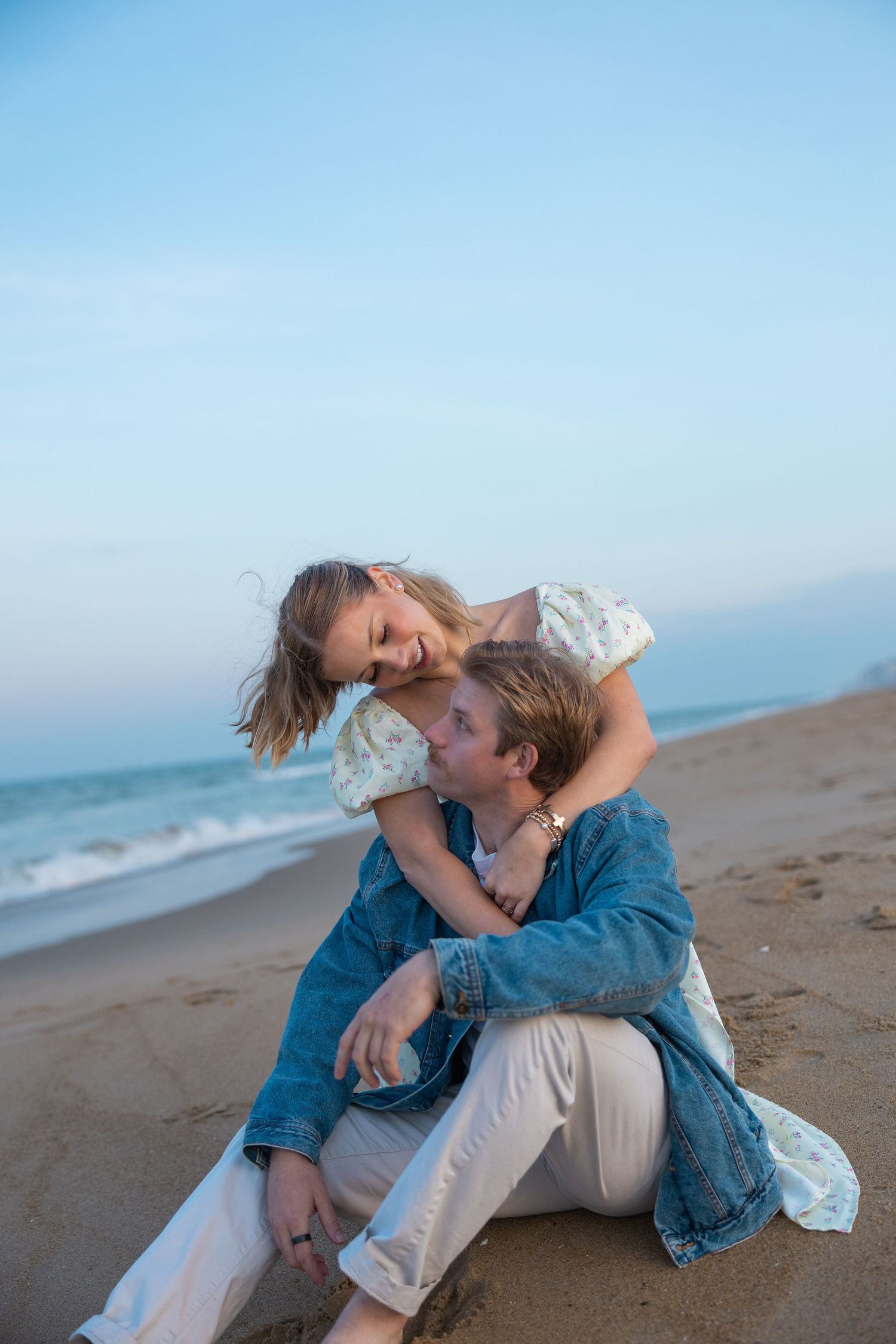 Romantic couple cuddling on sandy beach at dusk, woman leaning over smiling man.