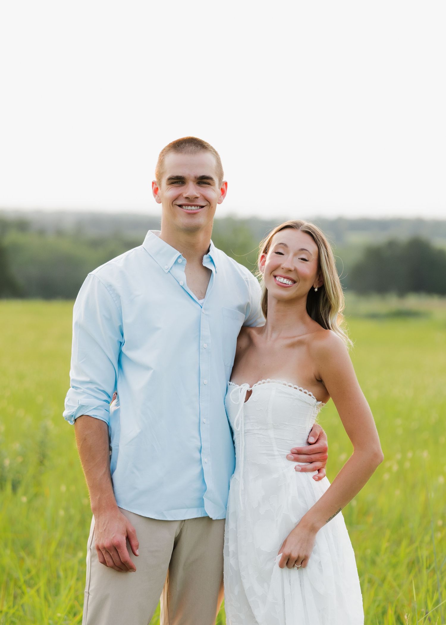 Smiling couple posing together in a green field, man in light blue shirt and woman in white strapless dress.