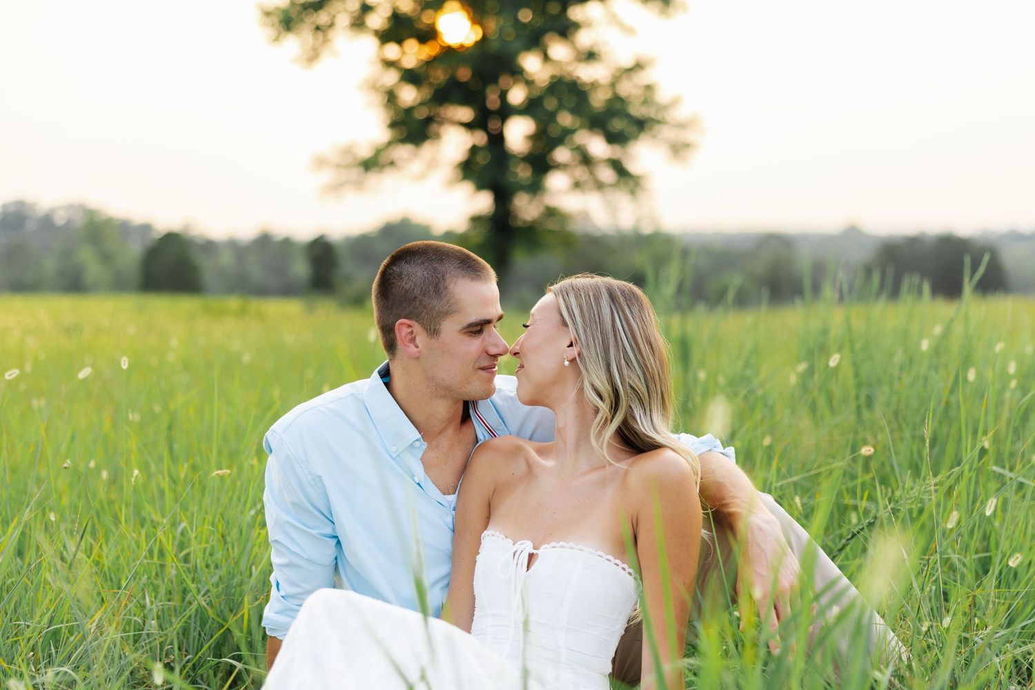 Couple sharing a romantic moment in a lush green field at sunset with a tree in the background.
