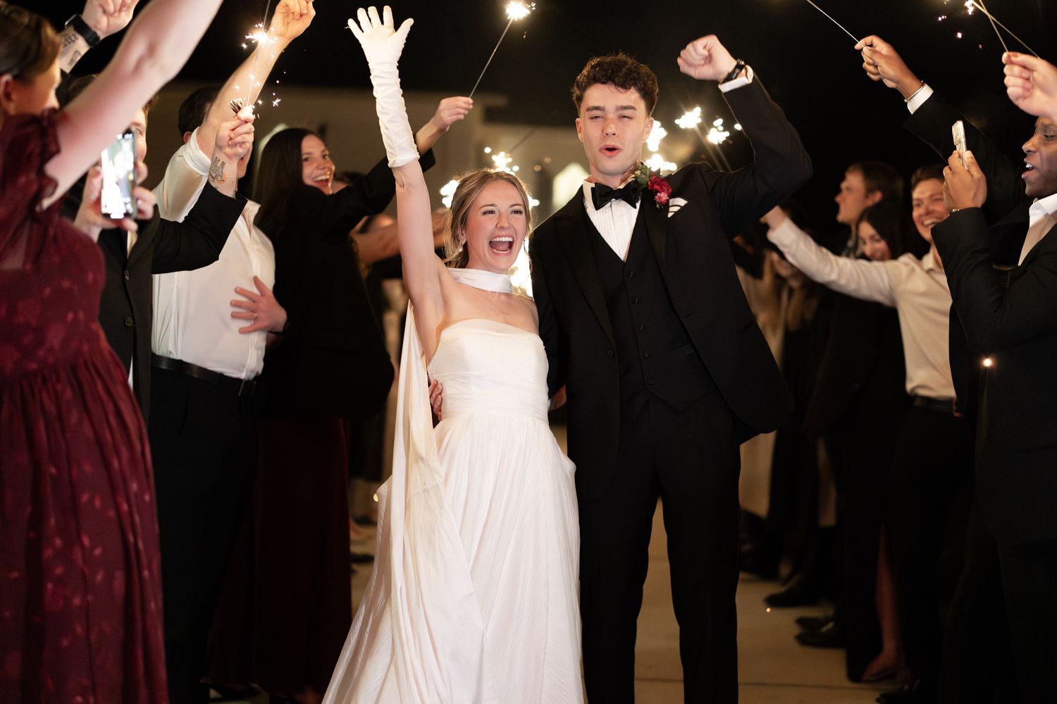 Joyful bride and groom walk through a sparkler tunnel exit, smiling and waving at their wedding celebration.