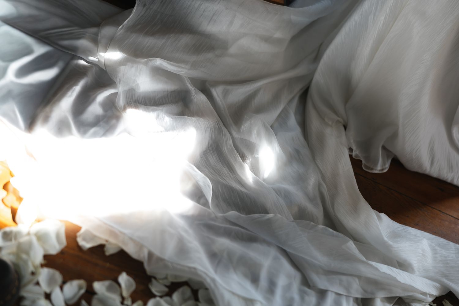 Close-up of flowing white wedding dress train with scattered flower petals on wooden floor.