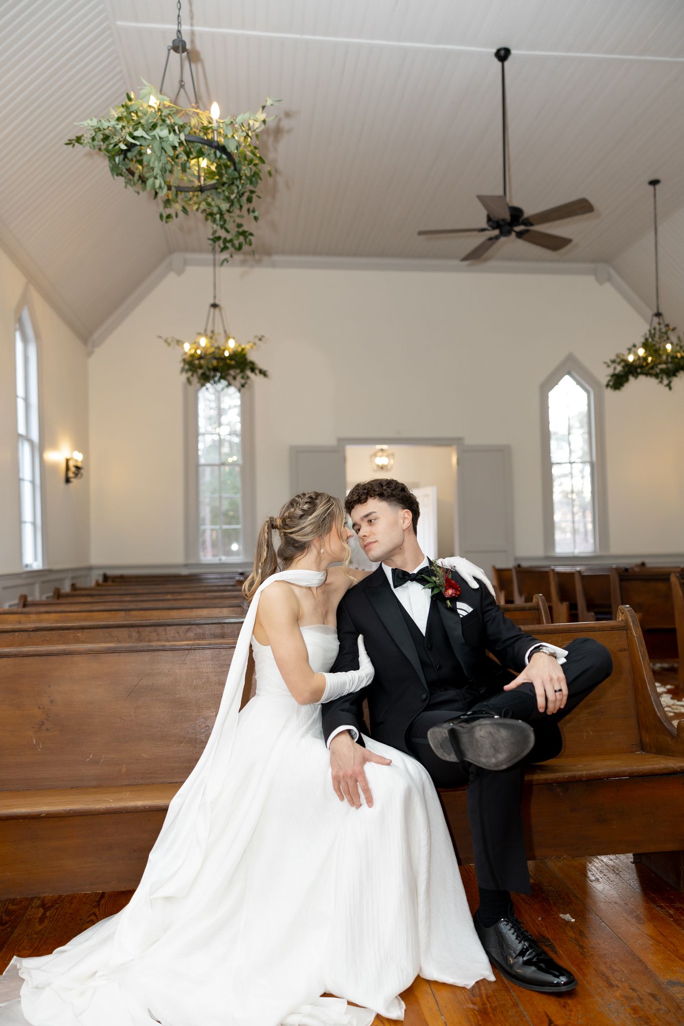Bride and groom sit closely on a church pew beneath hanging greenery chandeliers in a sunlit chapel.