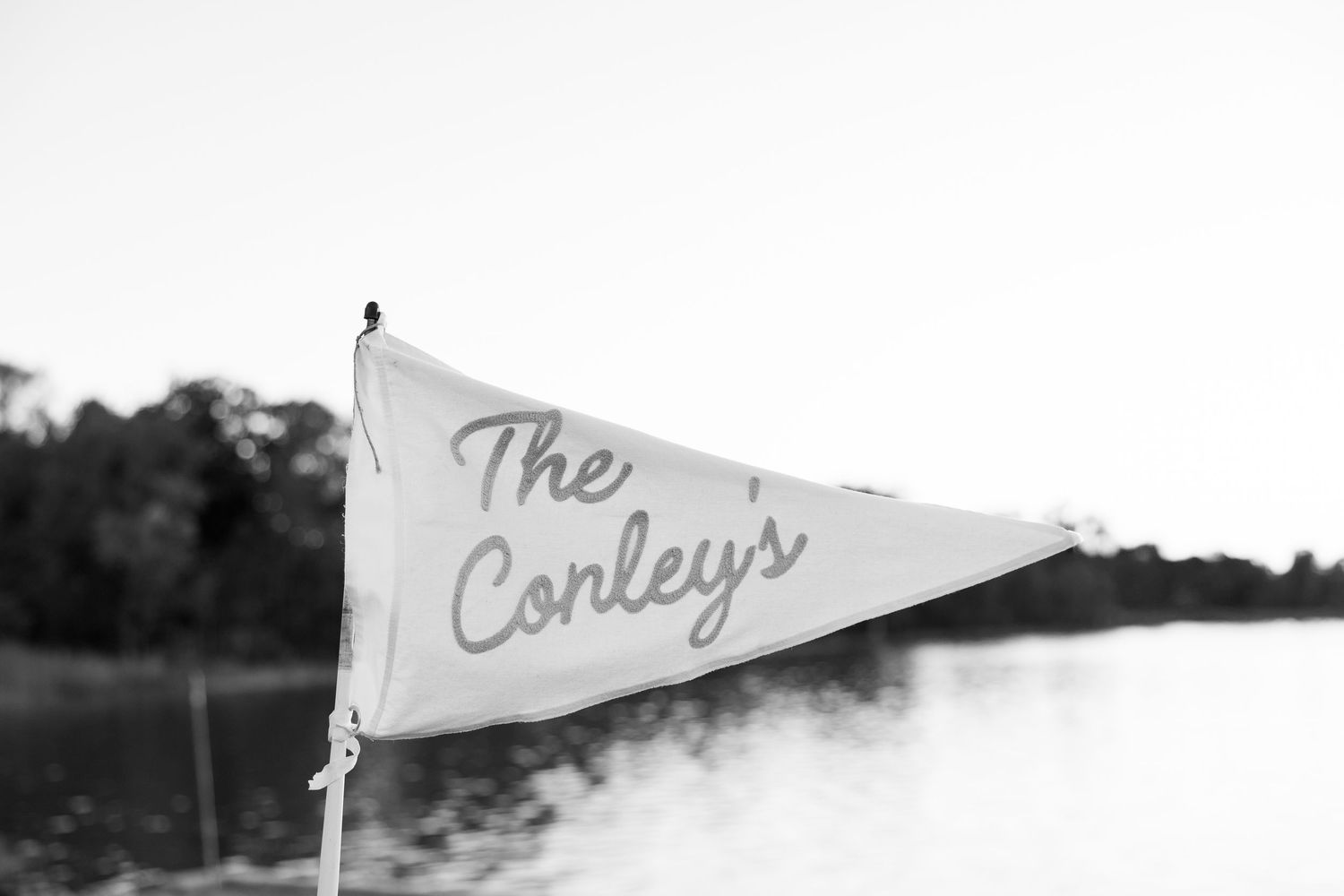 Black and white photo of a triangular pennant flag reading The Conleys near a lakeside with trees.