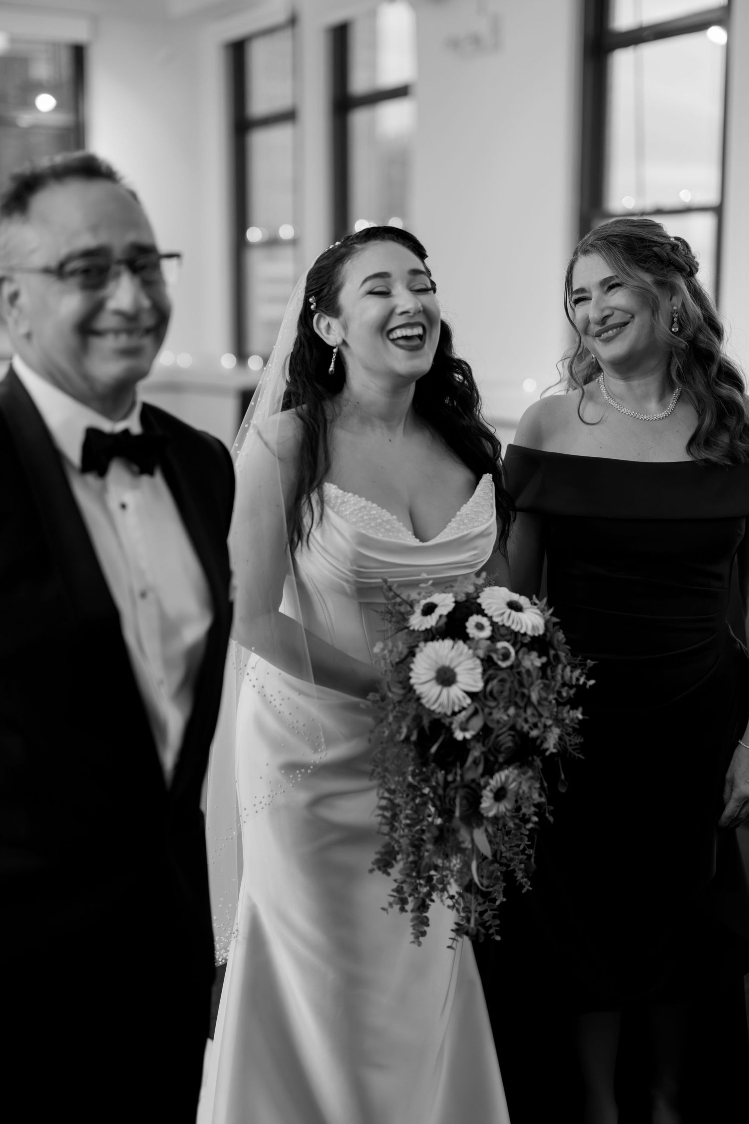 Black and white photo of a laughing bride in a satin gown holding a bouquet, flanked by smiling guests at a wedding.
