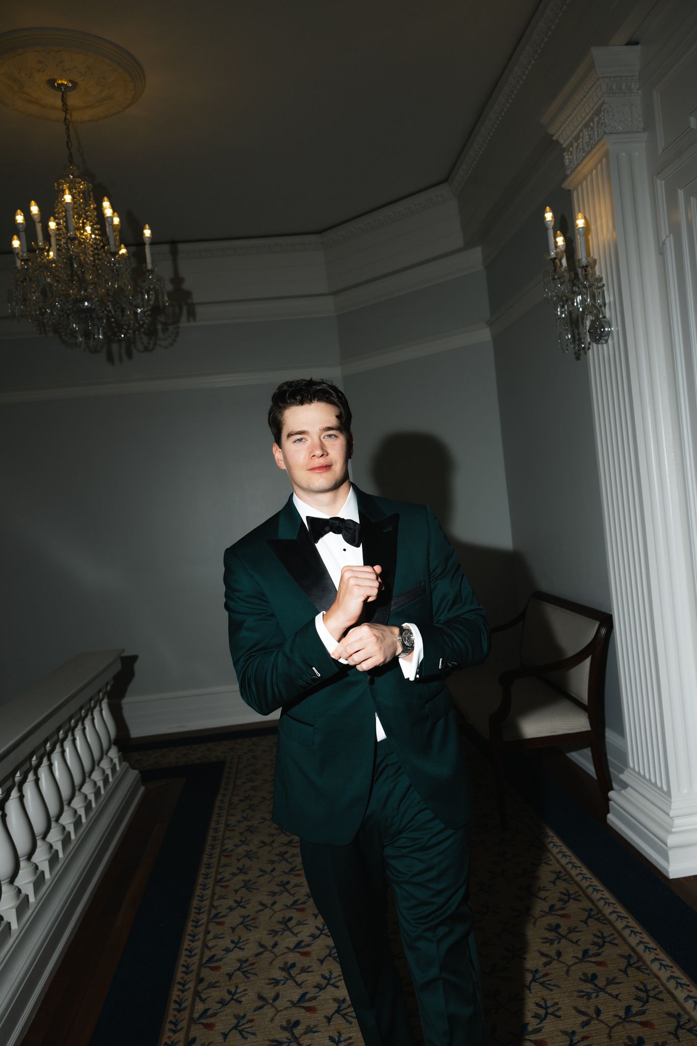 Elegant groom in green tuxedo adjusting cufflinks in ornate room with crystal chandeliers.