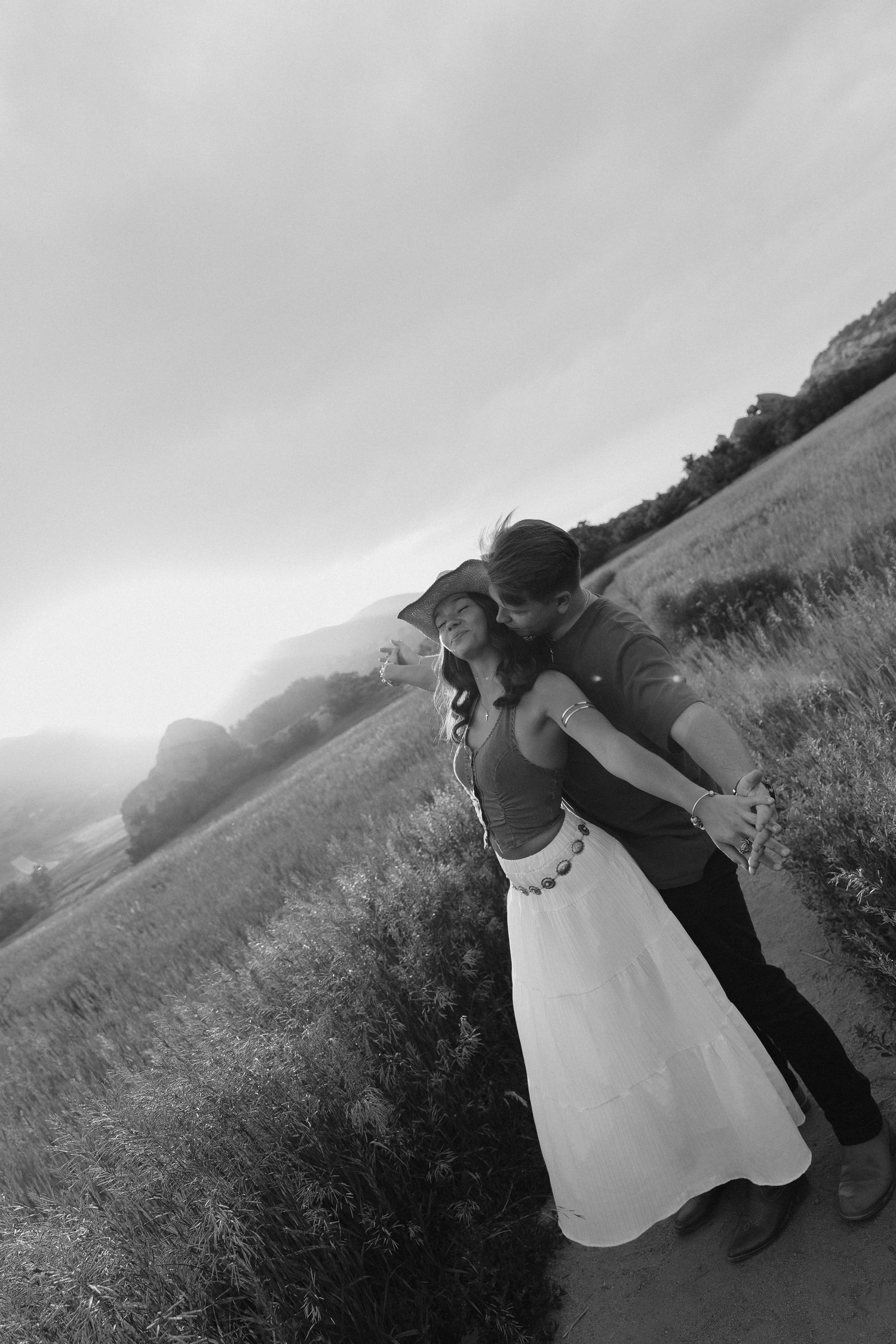 Black and white romantic photo of a couple embracing on a dirt path beside an open field at dusk.