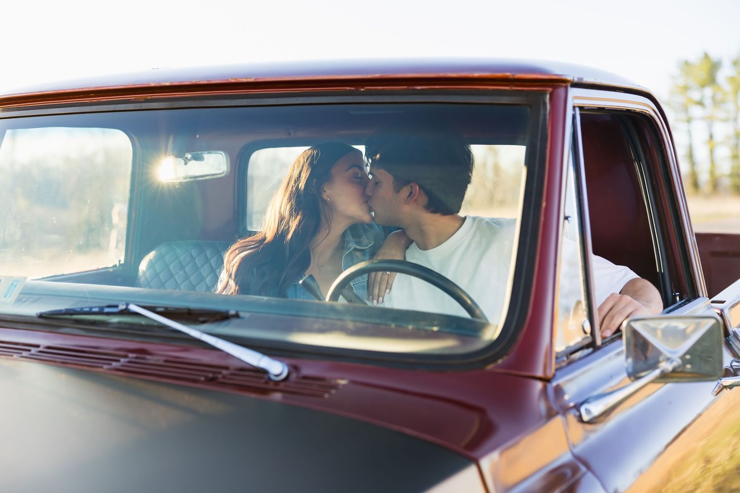 Couple sharing a romantic kiss inside a classic red pickup truck on a sunny day.